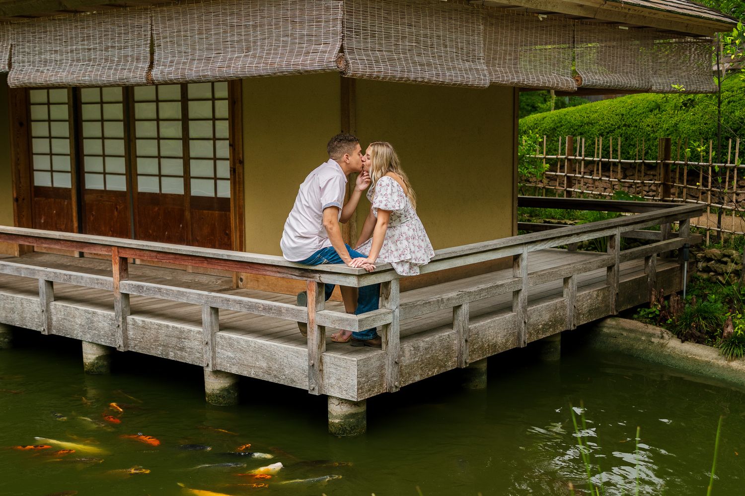 Couple sharing tender moment on wooden deck overlooking koi pond in Japanese garden setting.