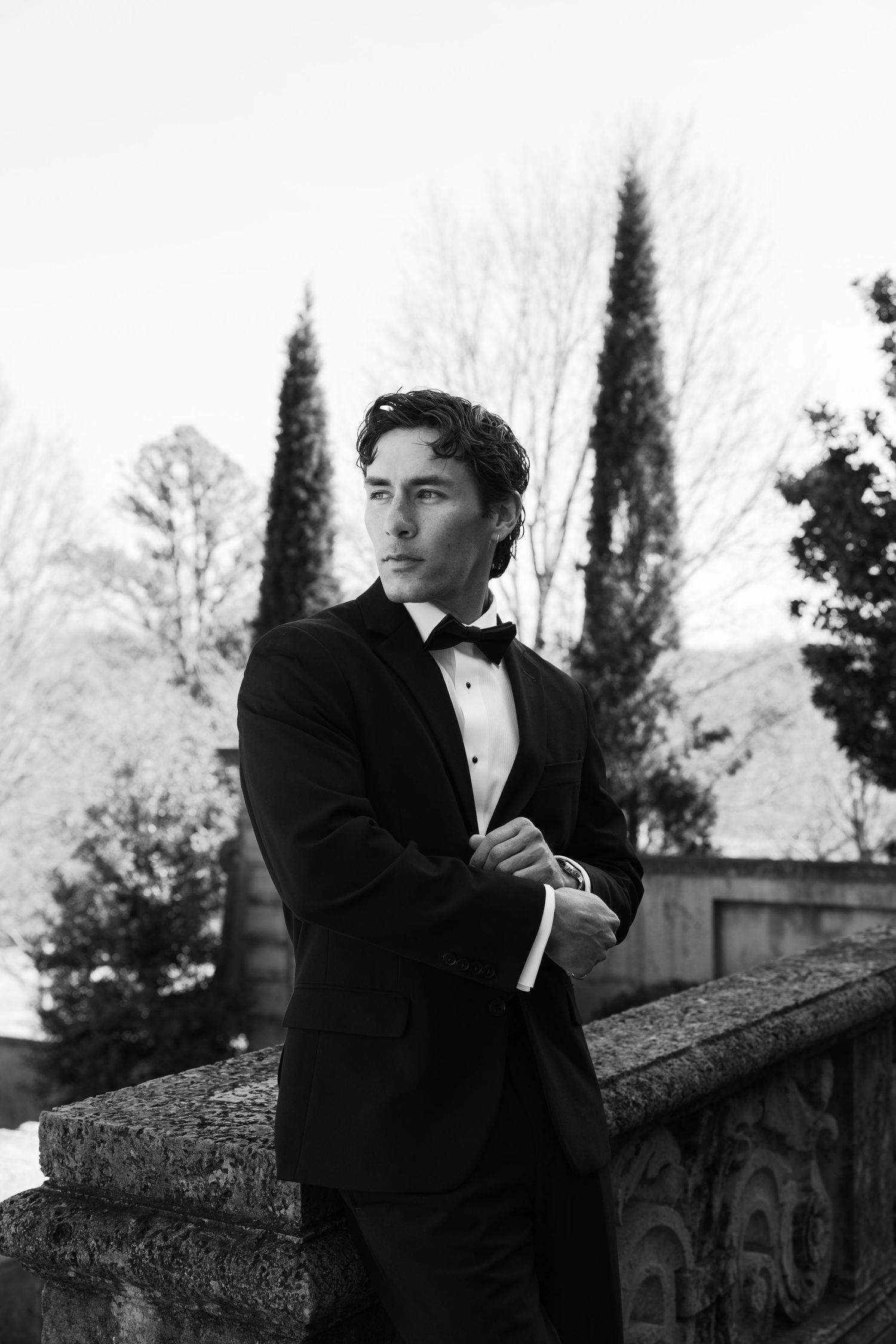 Elegant groom in black tuxedo and bow tie poses outdoors by stone balustrade with tall cypress trees in black and white.