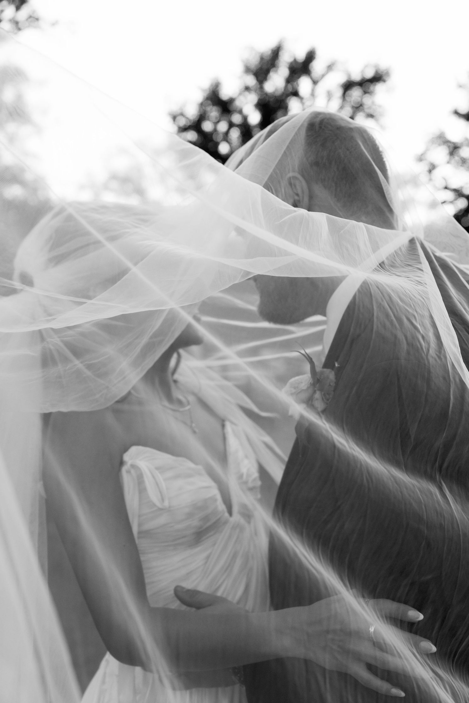Black and white photo of bride and groom sharing a kiss under a flowing wedding veil outdoors.