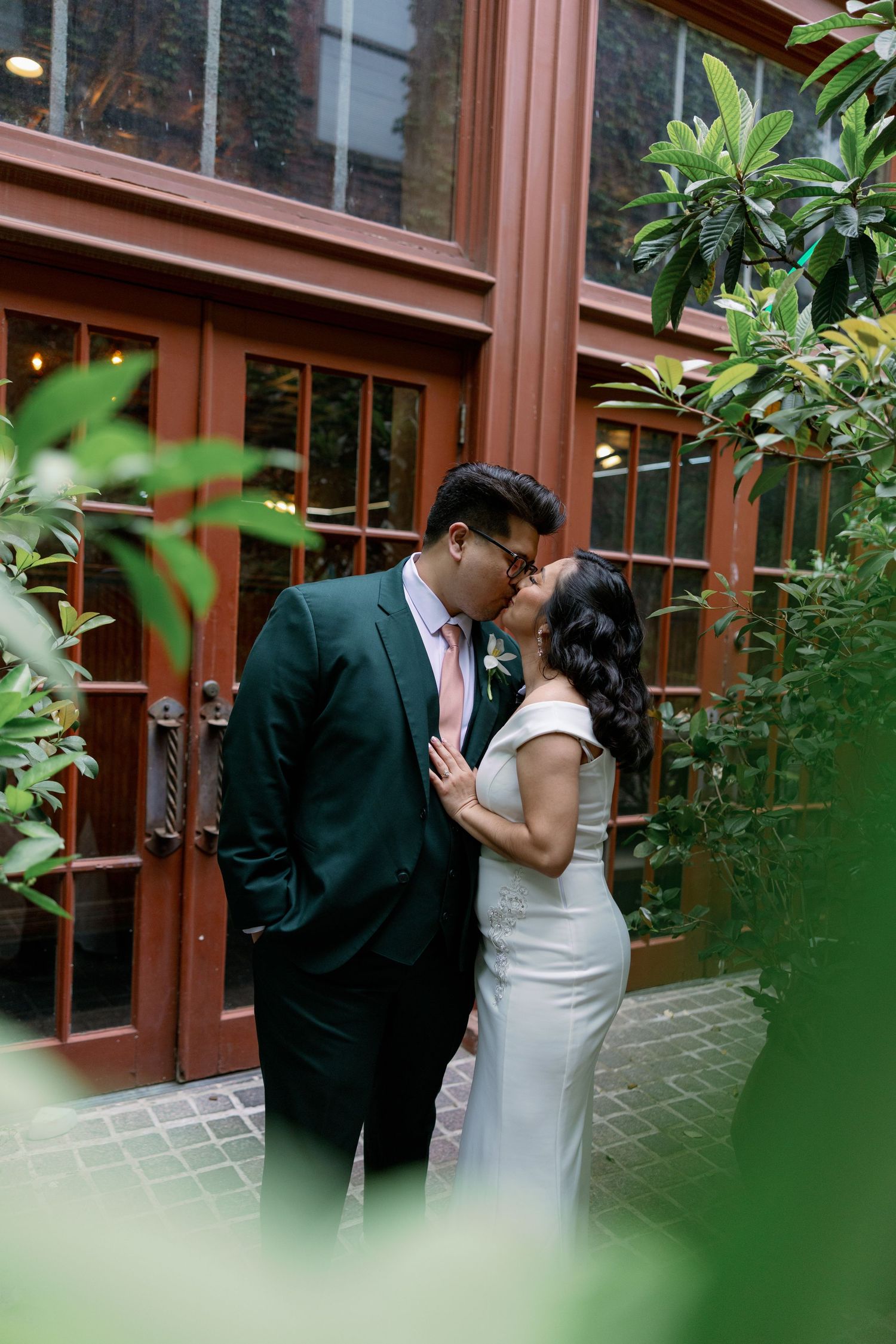 Couple sharing a romantic kiss in wedding attire, groom in dark green suit, bride in white dress, surrounded by lush greenery.