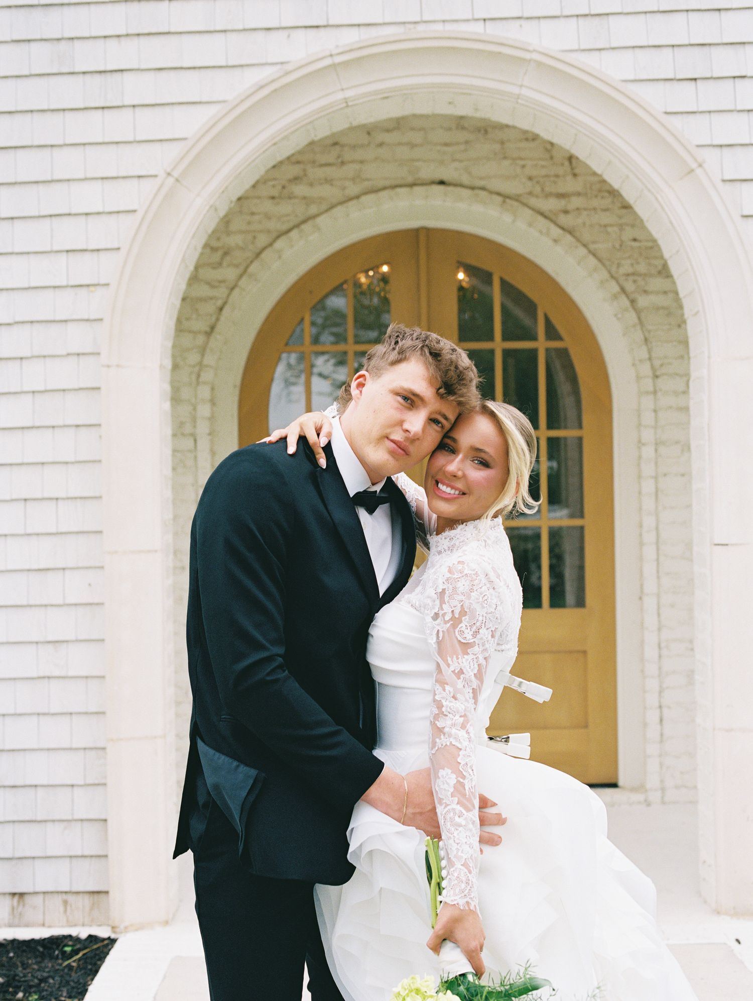 Bride and groom embrace in front of arched yellow door, wearing lace wedding gown and black tuxedo.