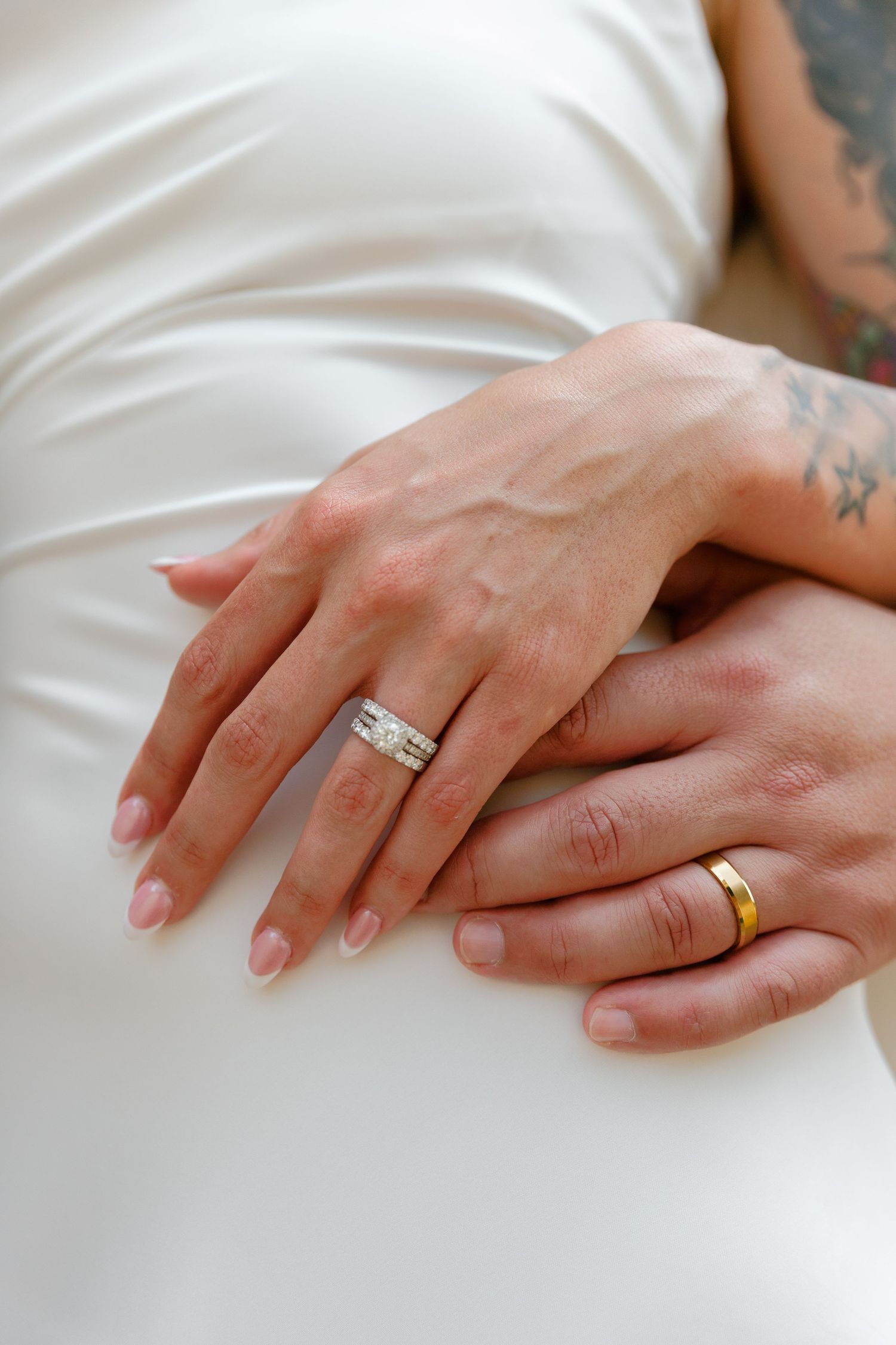 Two hands wearing wedding rings rest gently on a white dress showing both gold and diamond bands.