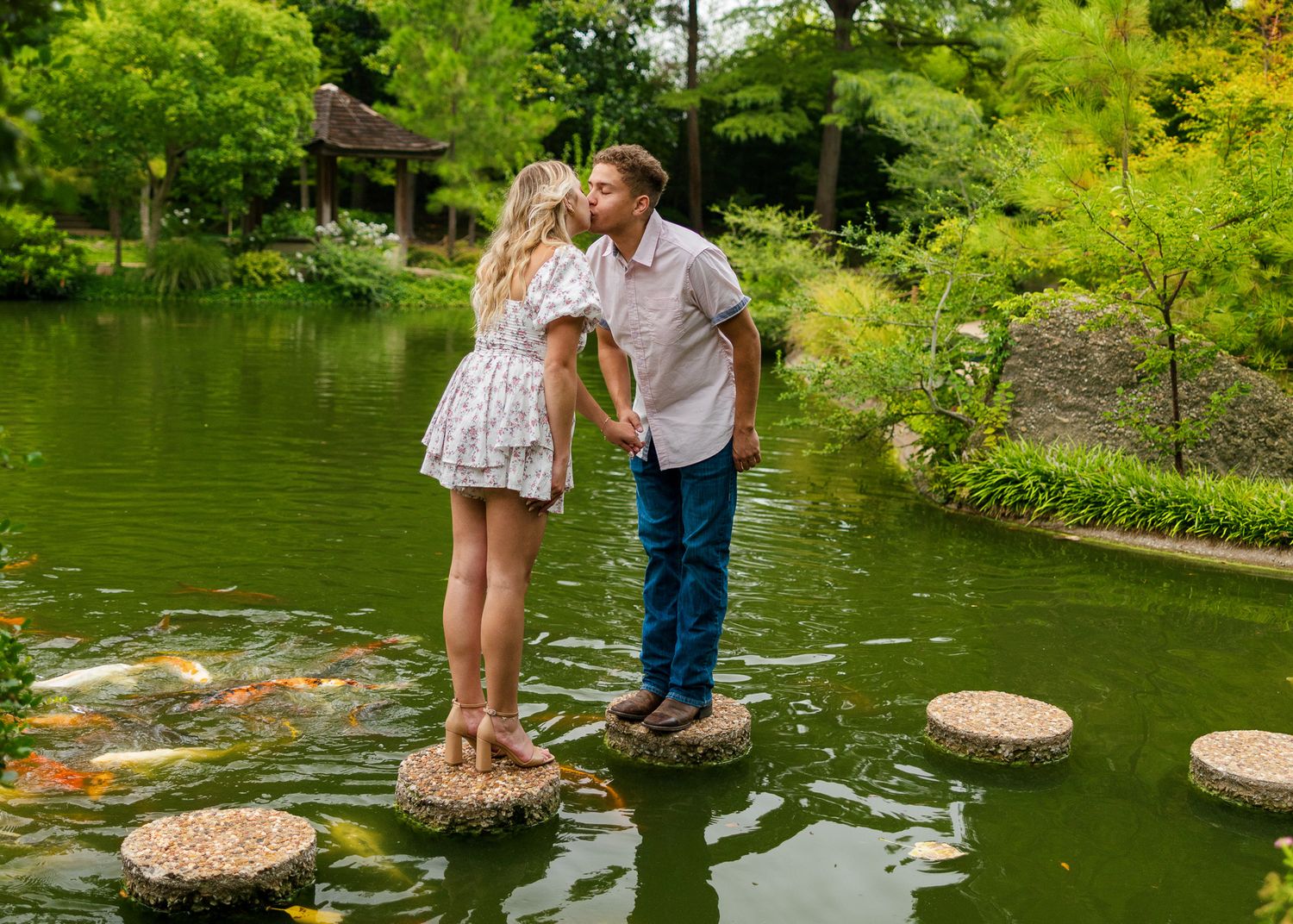 Two people stand on stepping stones across a serene green pond in a lush garden setting.