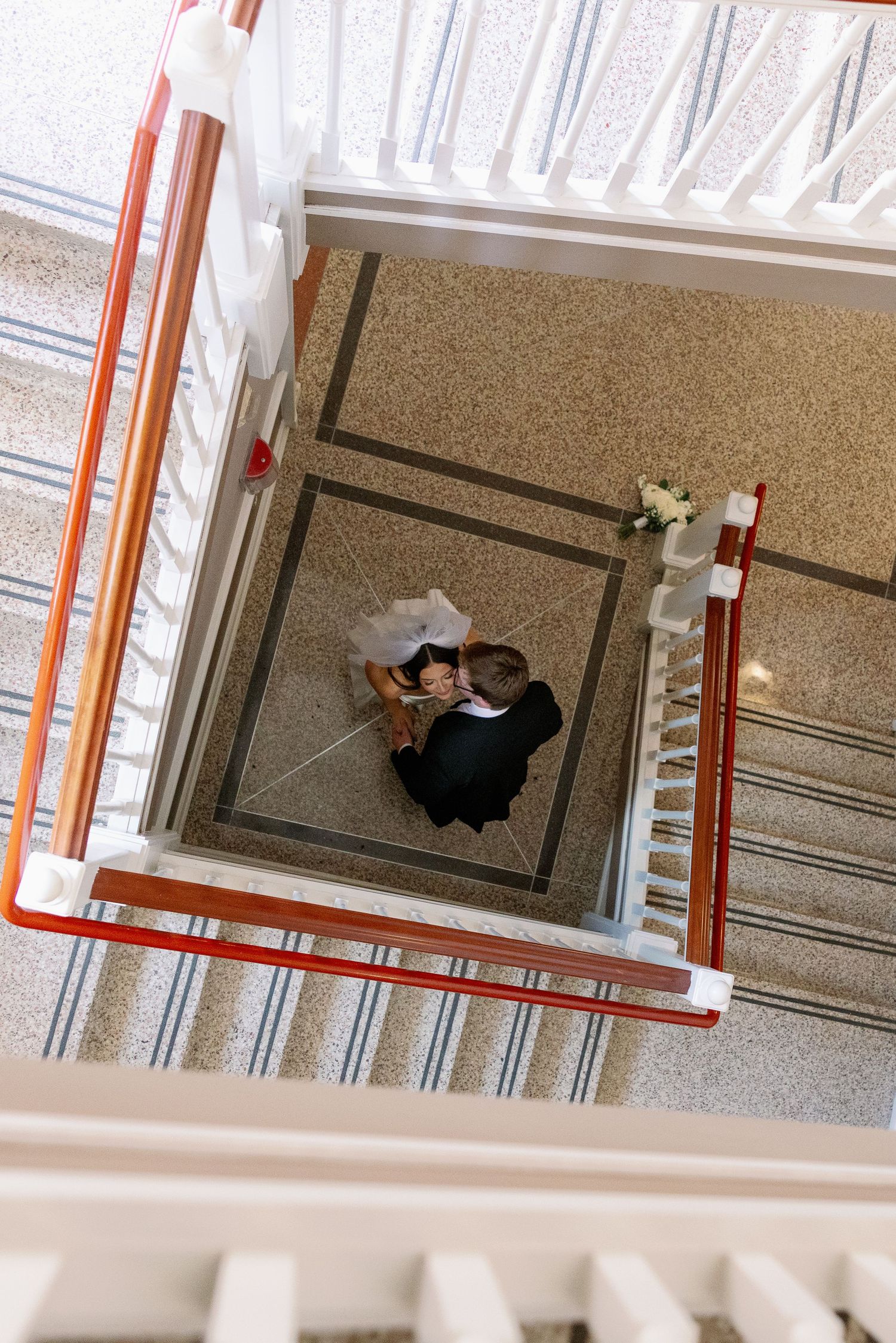 Aerial view of bride and groom kissing at the bottom of an elegant staircase with red railings.