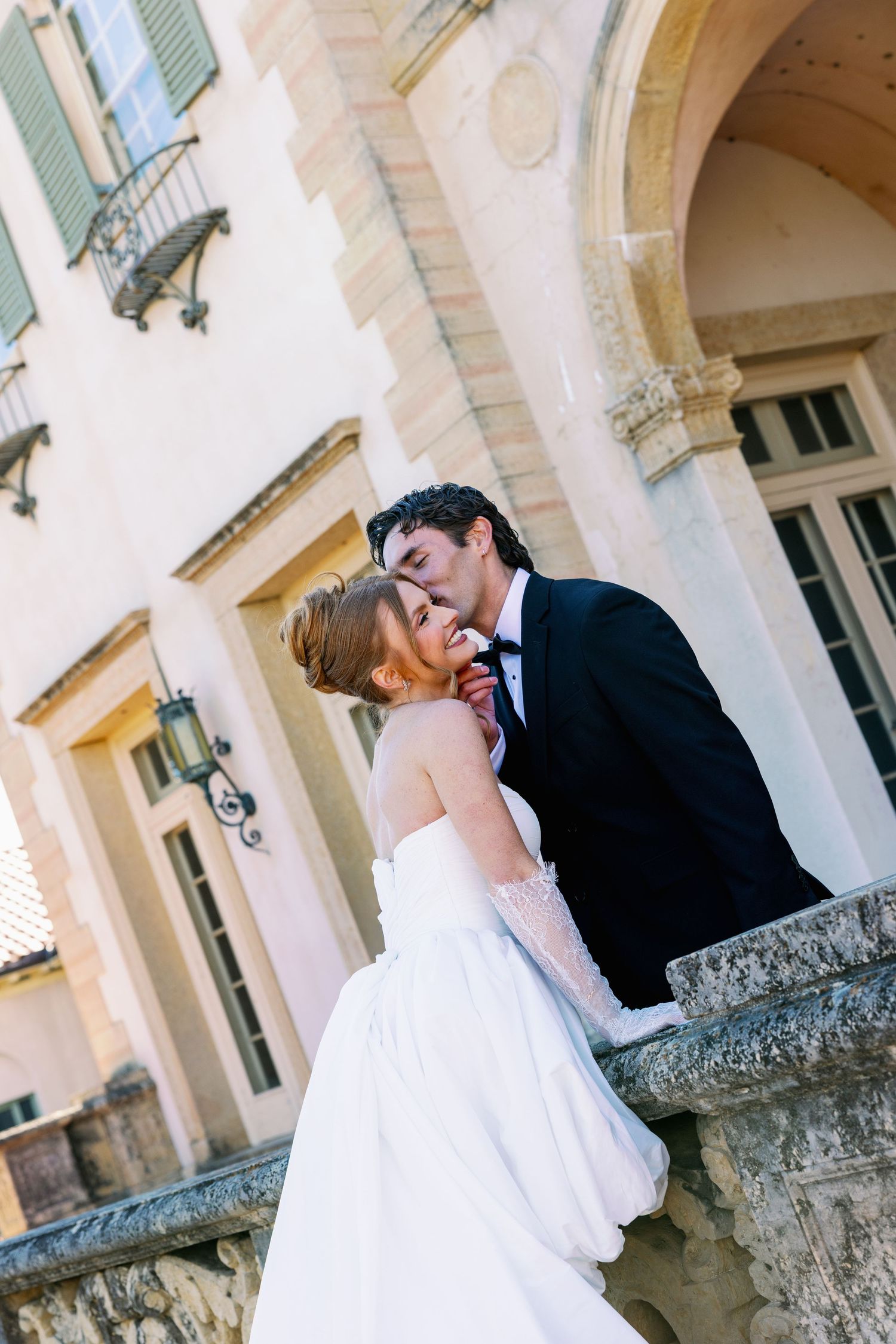 A bride and groom share a romantic kiss on a stone balcony outside an elegant historic building.