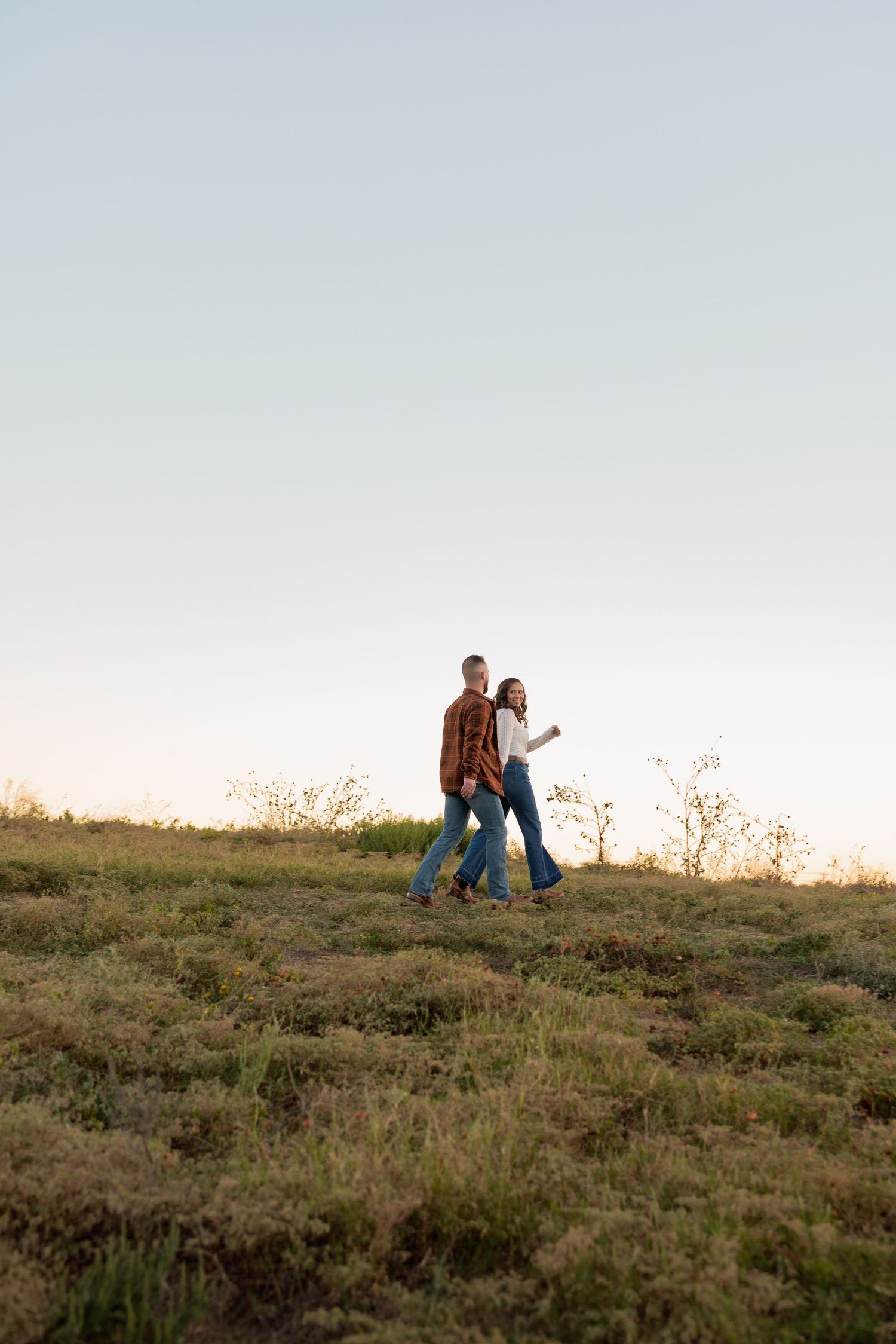Couple walking hand in hand through a grassy field at golden hour, wearing jeans and casual tops.
