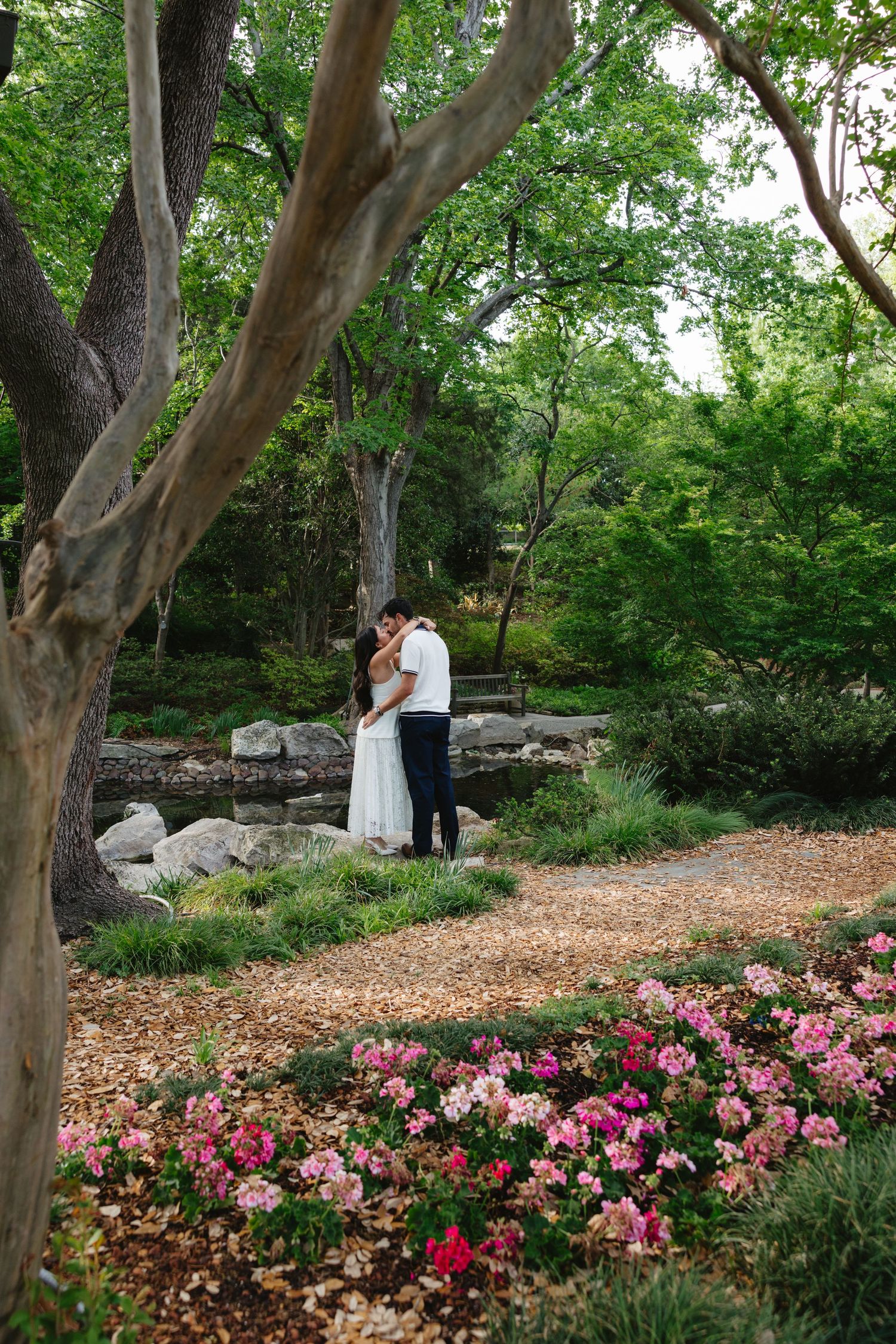 Couple embracing in a lush garden surrounded by pink flowers, green trees, and rocky landscape on a romantic outdoor occasion.