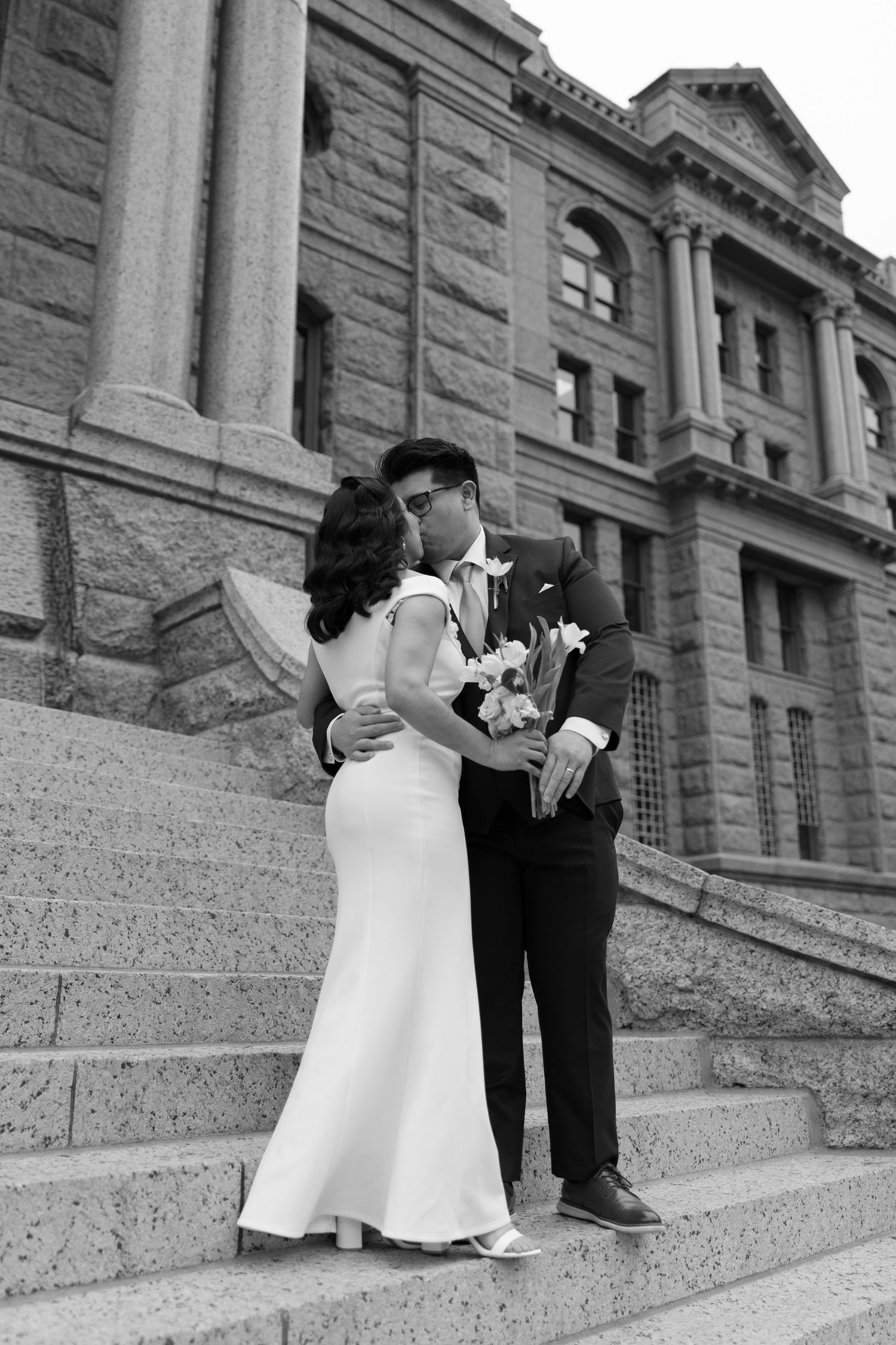 Black and white photo of a couple kissing on stone steps outside a grand building, bride in white gown holding bouquet.