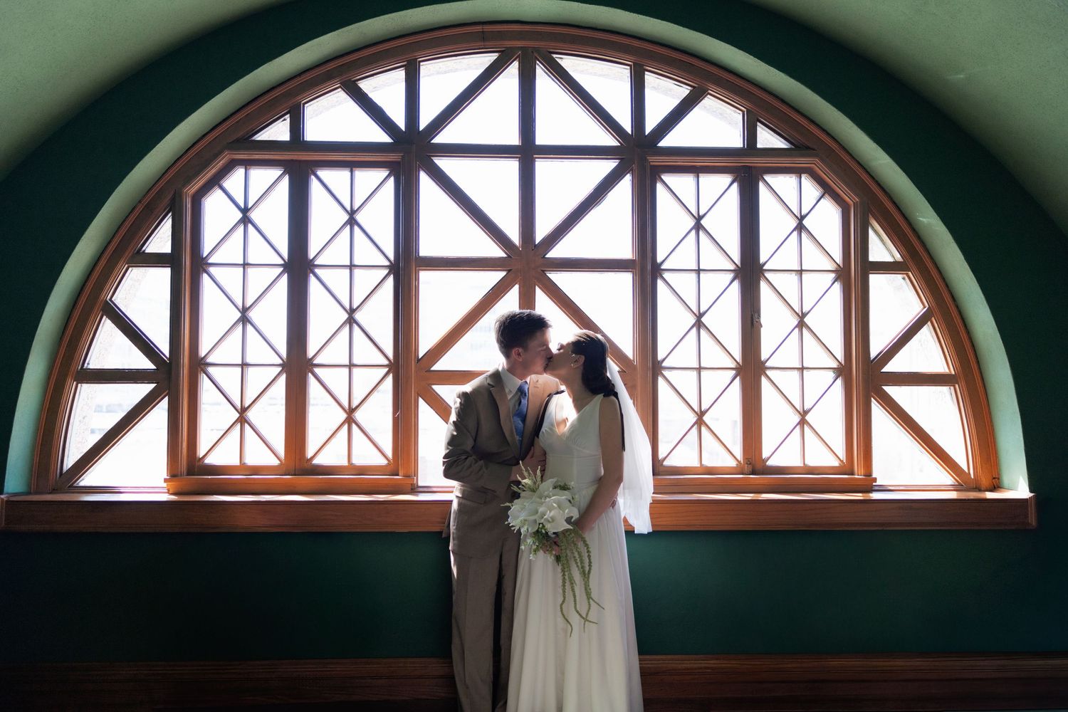 Bride and groom sharing a romantic kiss silhouetted against a large arched window with geometric panes.