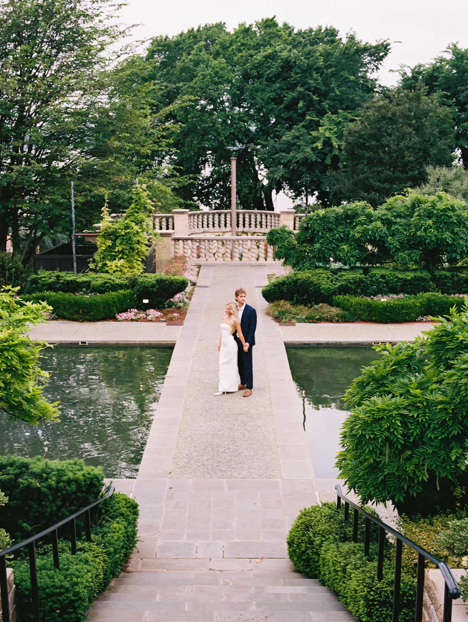 Bride and groom embrace on a stone pathway flanked by manicured hedges and reflecting pools in an elegant garden venue.