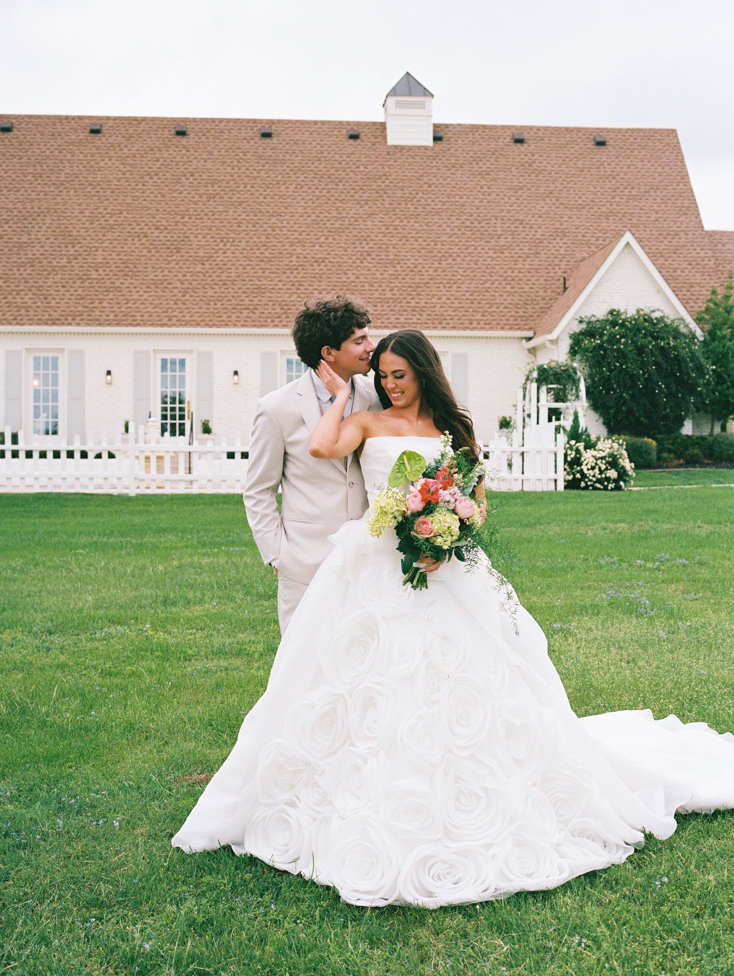 A bride in a strapless ball gown with a floral bouquet stands with a groom in a tan suit outside a white venue.