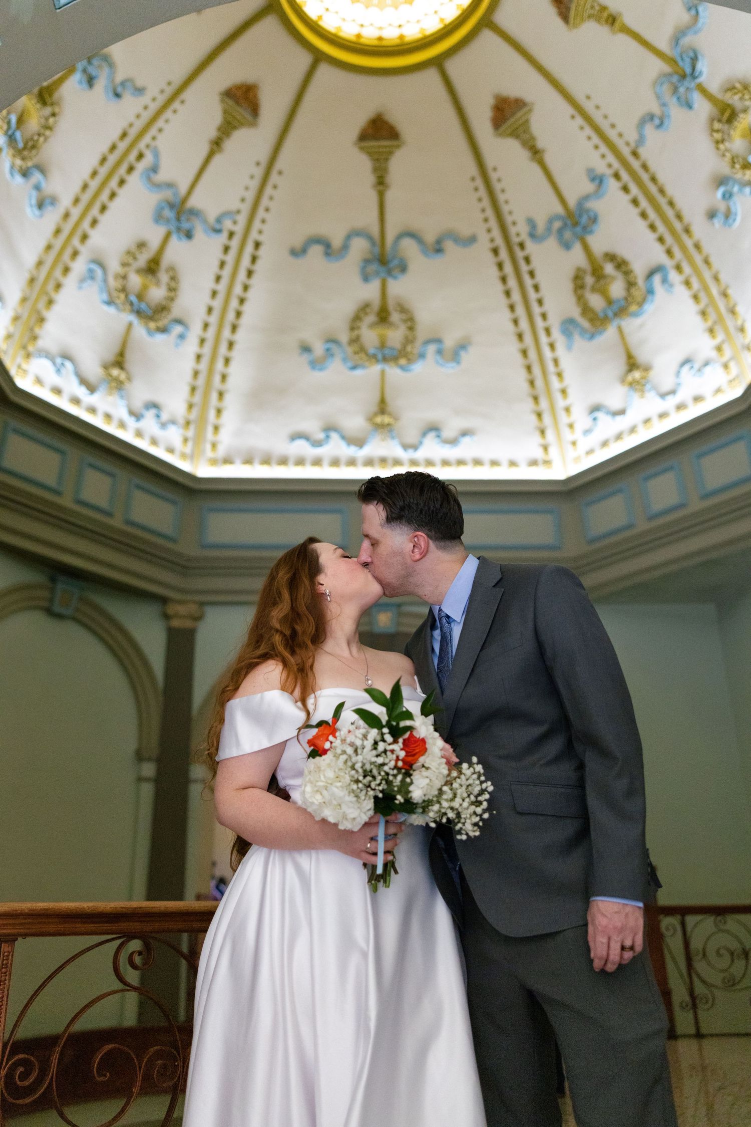 Bride and groom share a kiss beneath an ornate painted dome ceiling in an elegant wedding venue.
