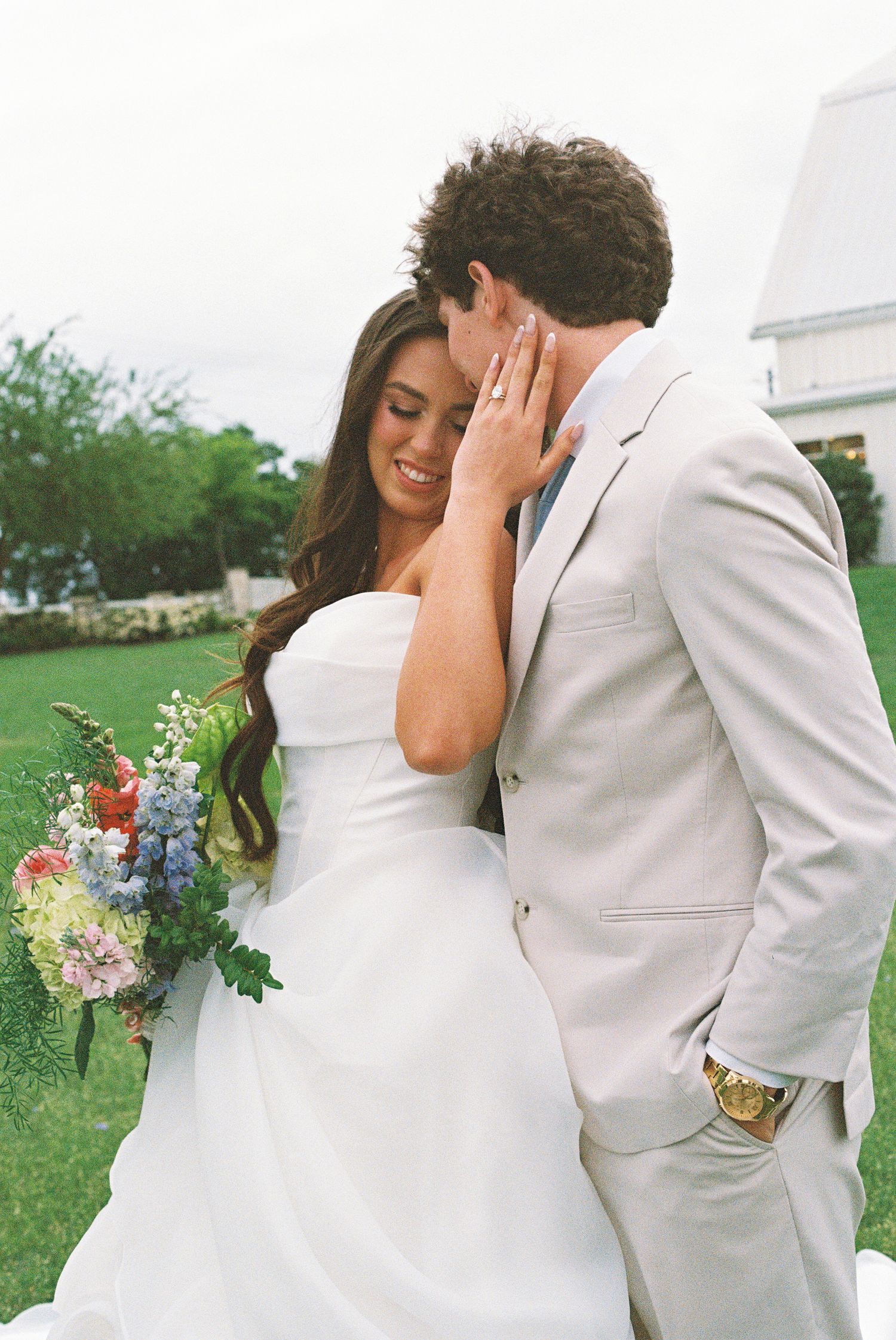 Bride in strapless white gown holding wildflower bouquet embraces groom in tan suit outdoors at wedding.