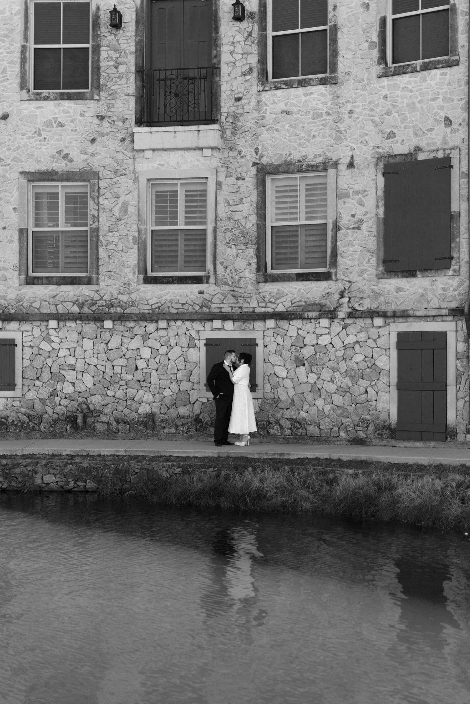 Black and white photo of a couple embracing by a canal in front of a rustic stone building with shuttered windows.