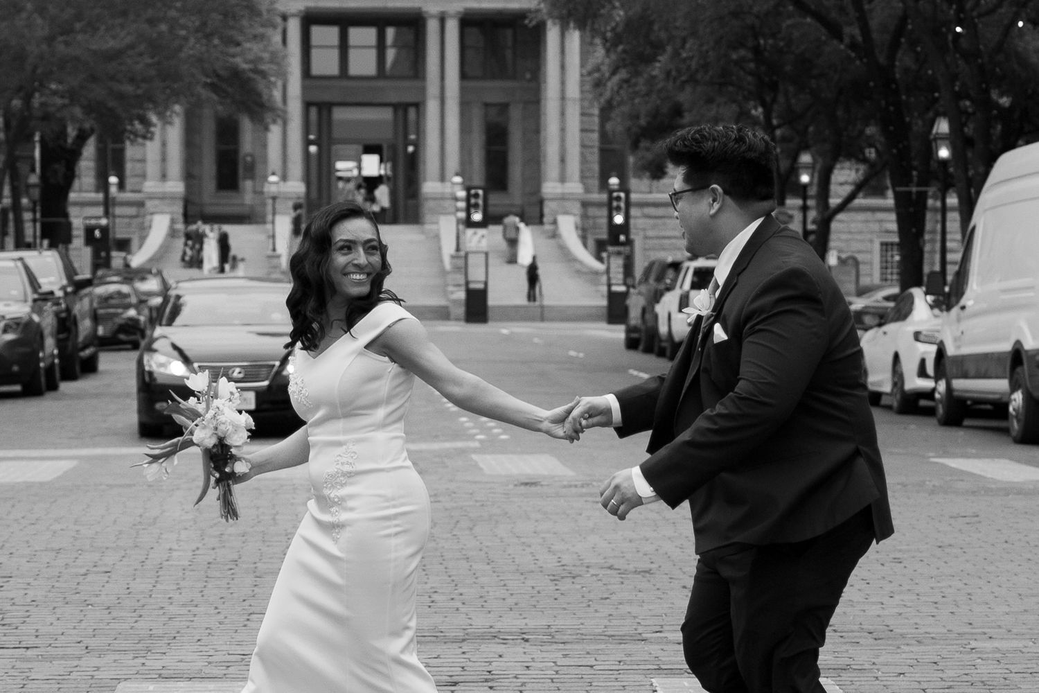 Black and white photo of a bride and groom holding hands, walking joyfully on a city street on their wedding day.