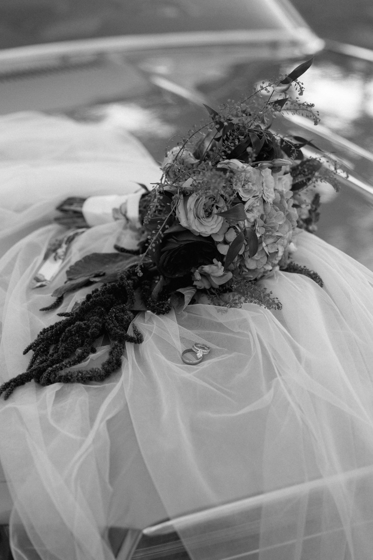 Black and white photo of a bridal bouquet with roses and greenery resting on a wedding veil with rings on a classic car.