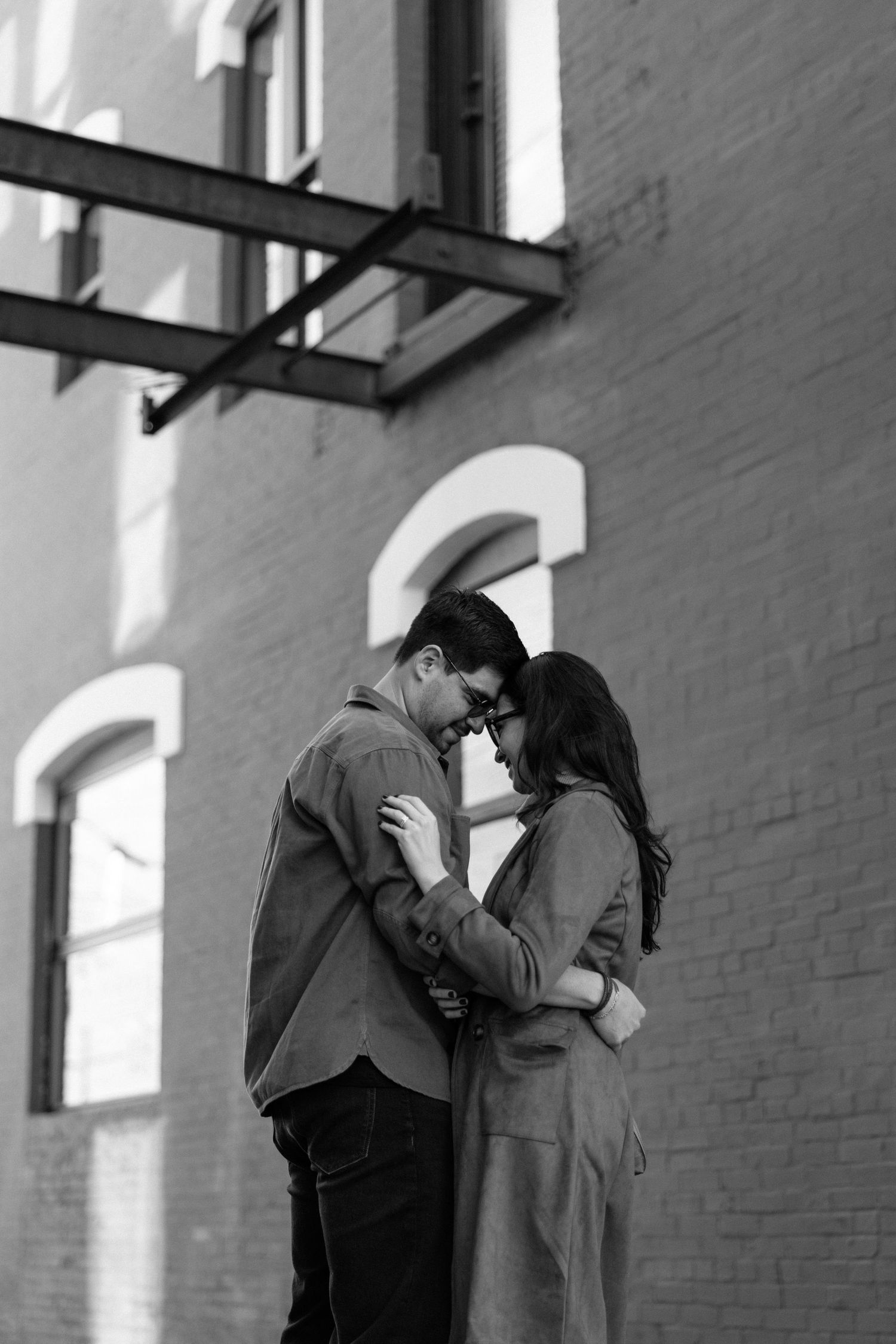 Black and white photo of a couple embracing tenderly against a brick building wall outdoors.