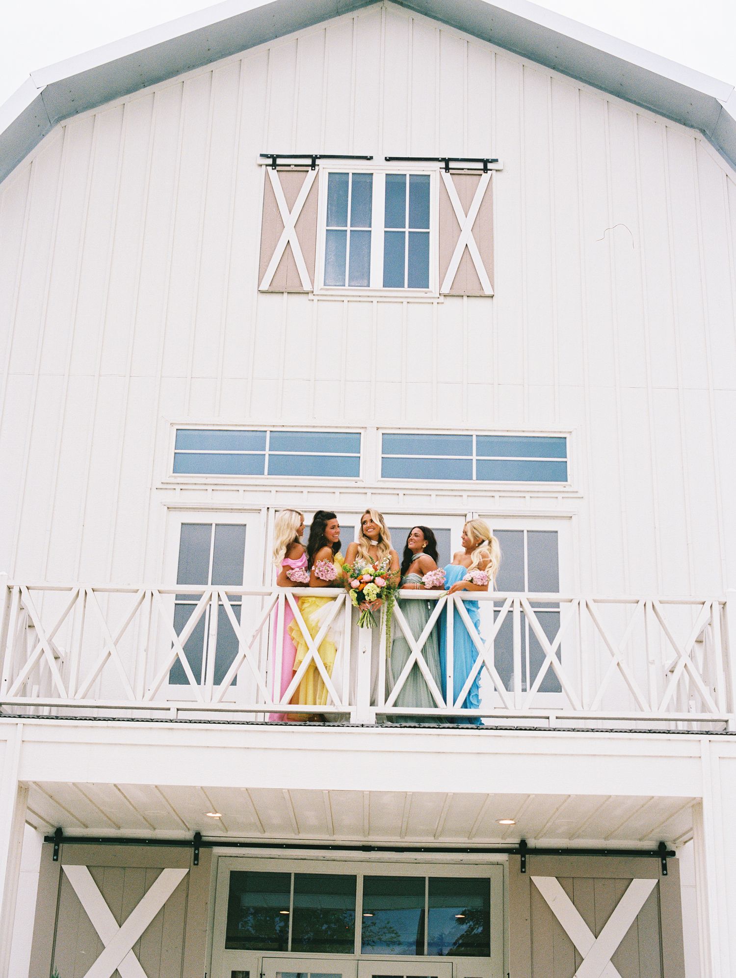 Bridesmaids in colorful dresses stand on a white barn balcony with a bride holding a bouquet.