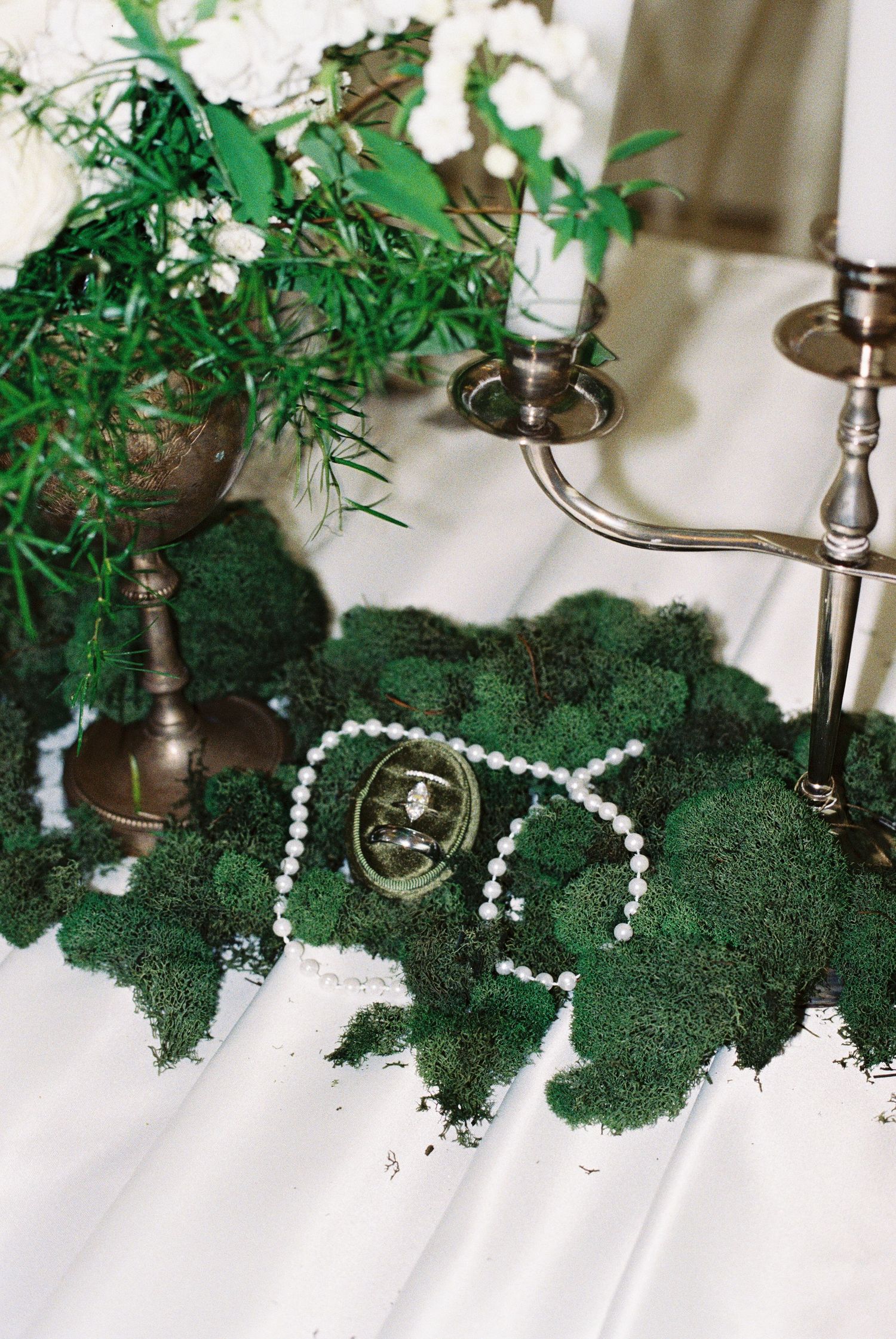 Wedding rings and pearl jewelry displayed on lush green moss with silver candelabra and white floral arrangement.