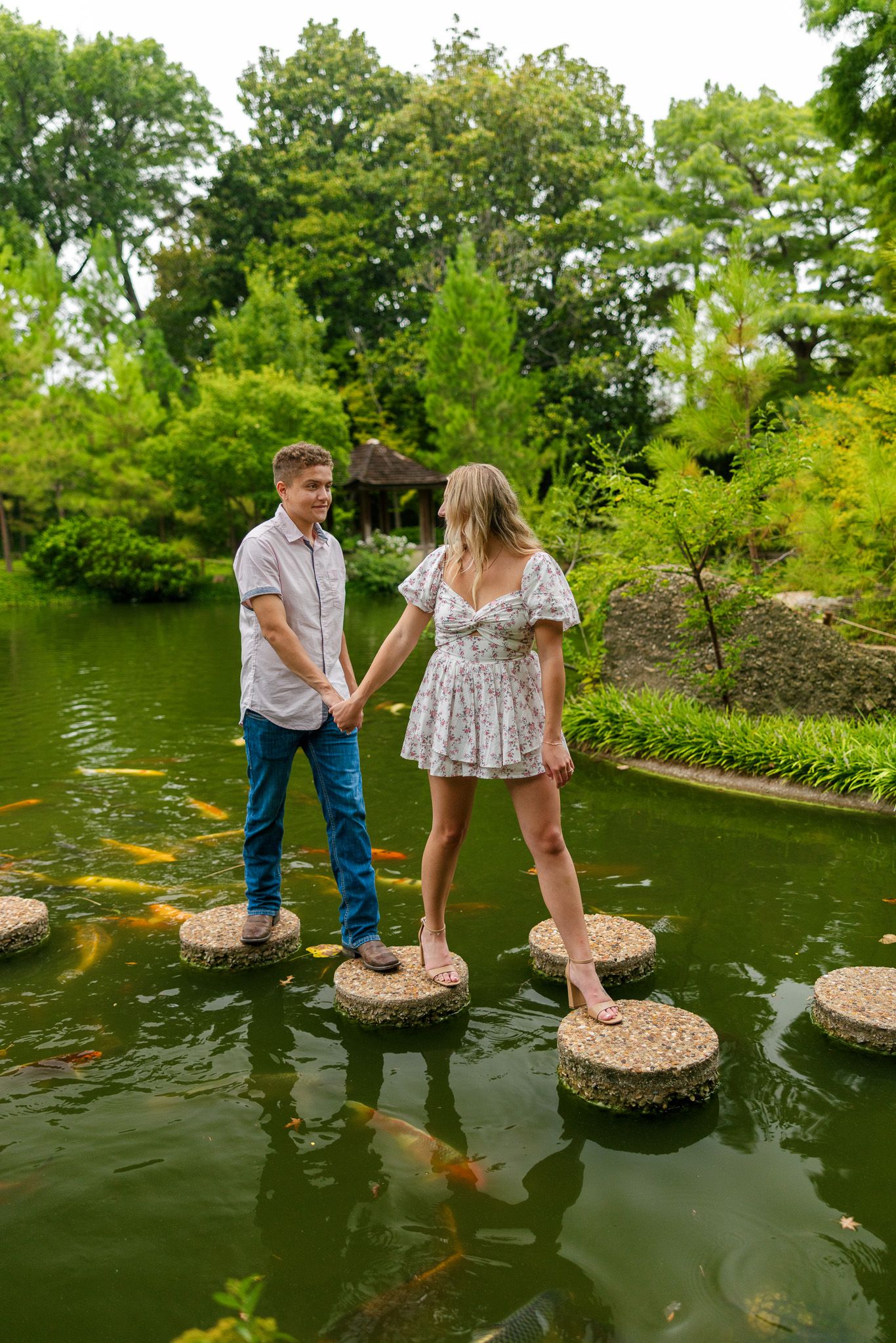 A couple playfully balances on stepping stones across a serene pond surrounded by lush greenery.