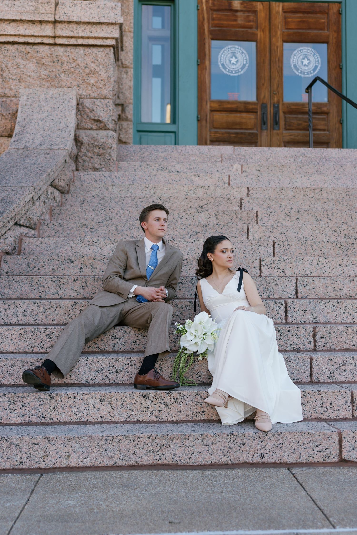 Bride and groom sitting on granite steps of a historic building, bride holding white bouquet in wedding attire.
