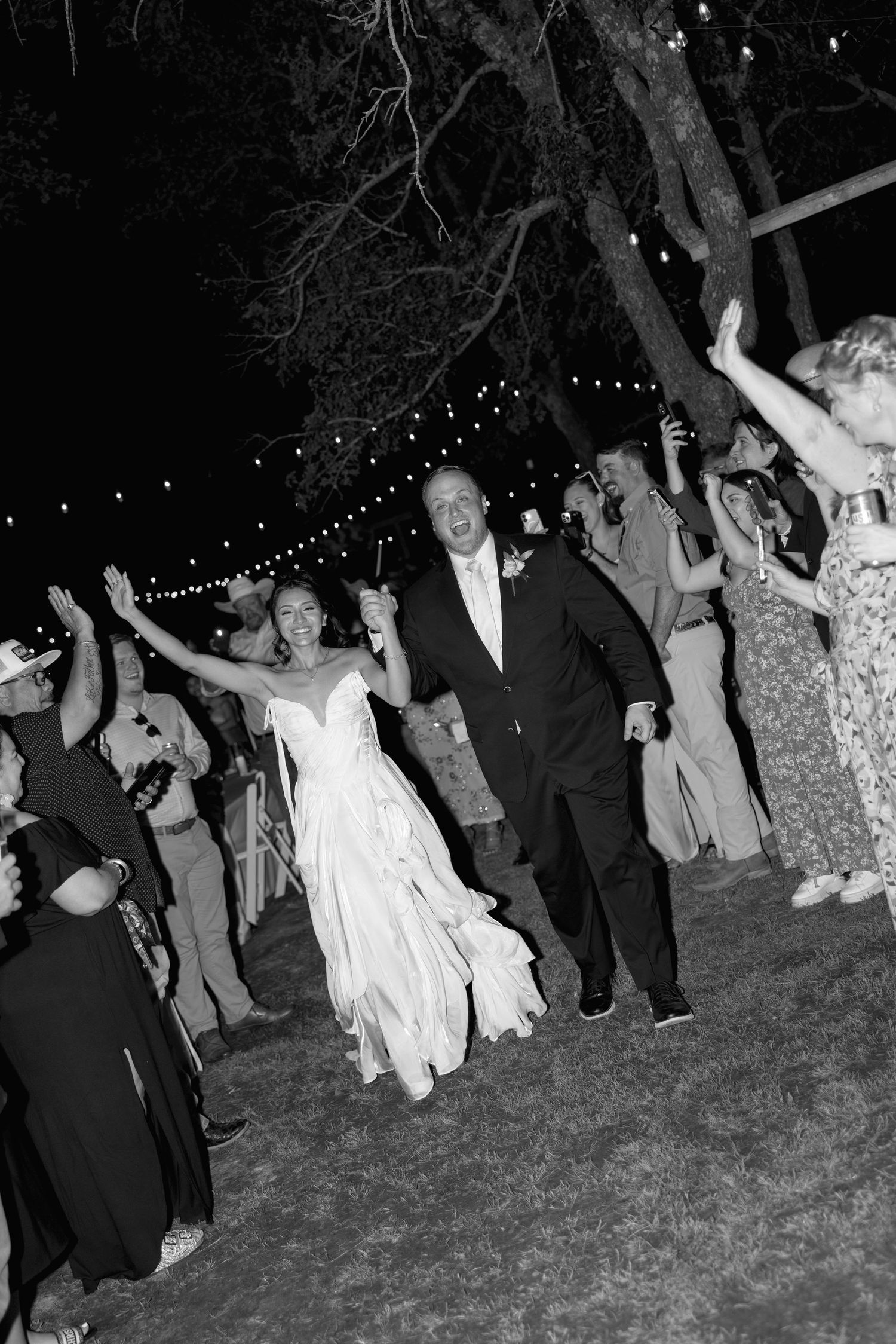 Bride and groom celebrate joyfully with cheering guests during outdoor wedding reception at night.