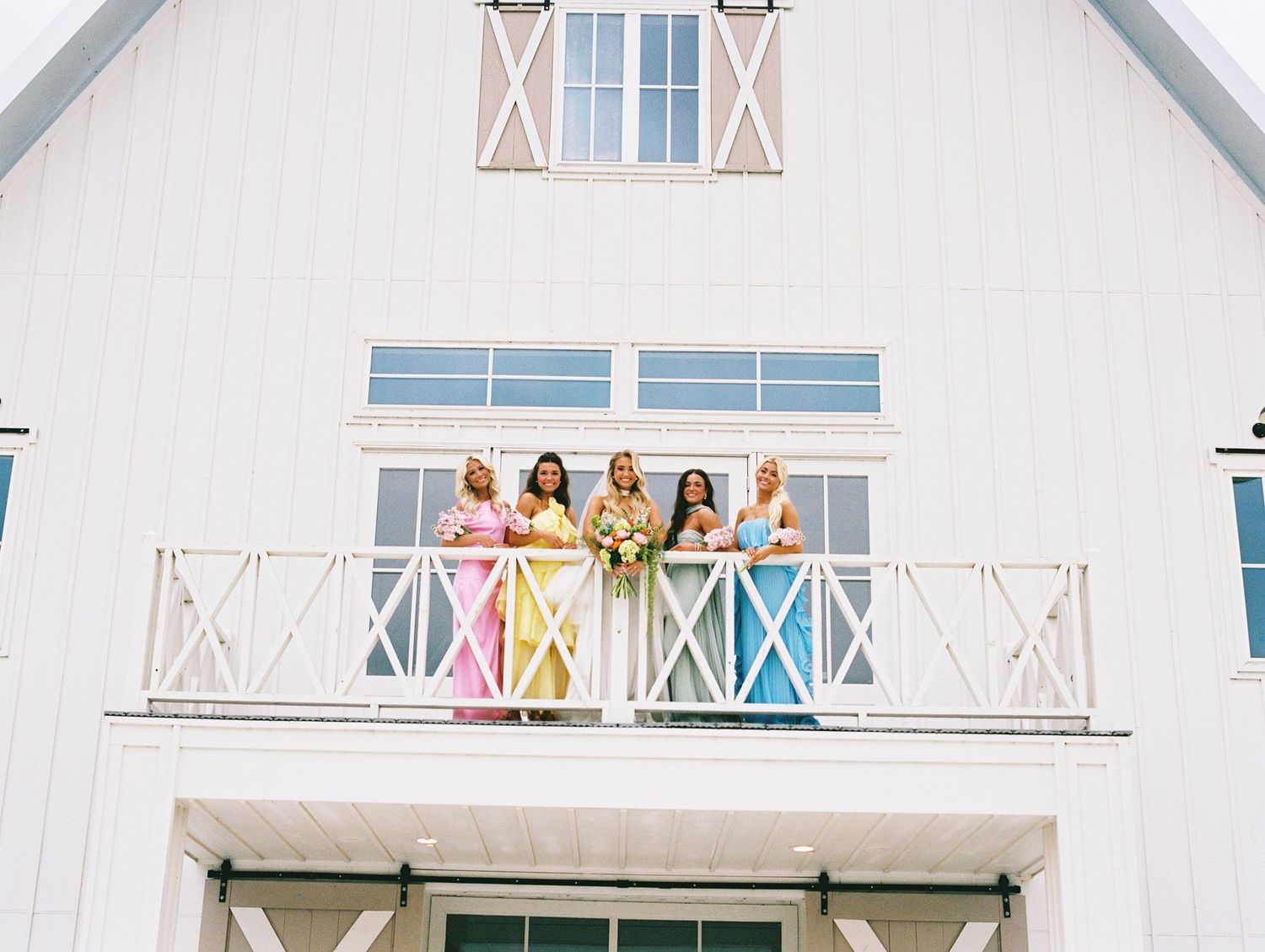 Bridesmaids in colorful dresses stand on a white barn balcony with a bride holding a bouquet.
