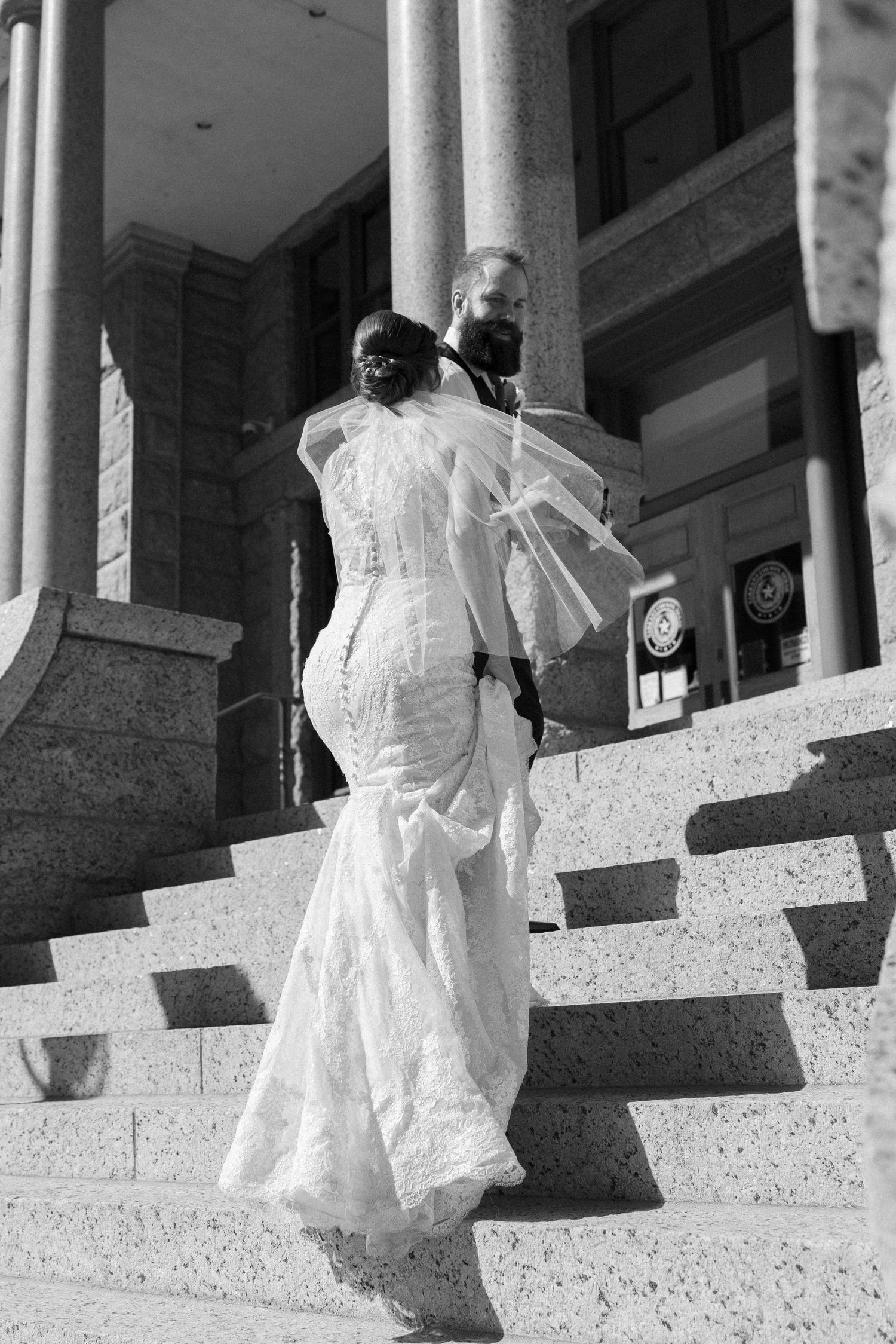 Black and white photo of a bride in lace gown and groom walking up stone steps of a grand building.