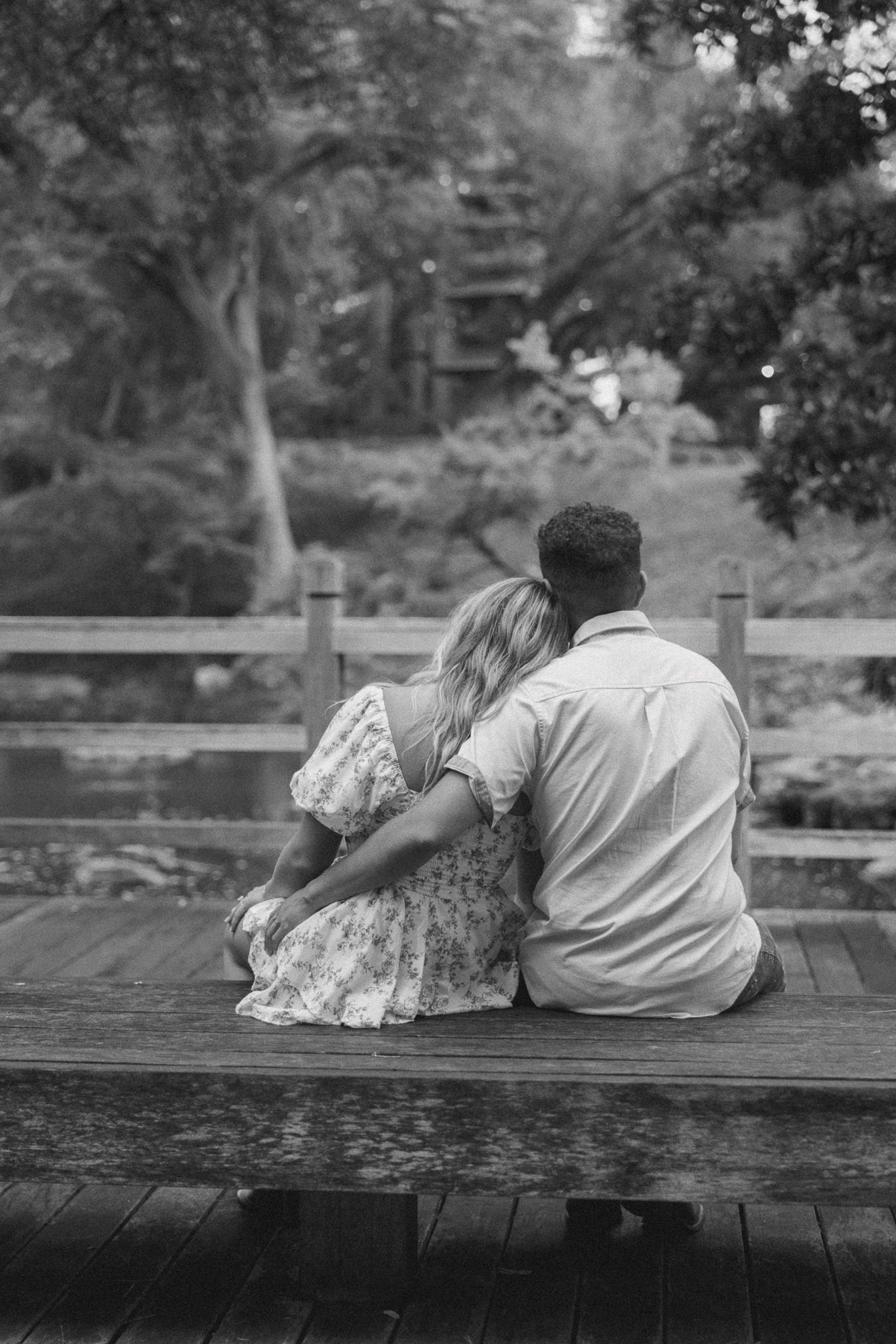 A couple sits close together on a wooden dock overlooking a peaceful pond in a serene garden setting.