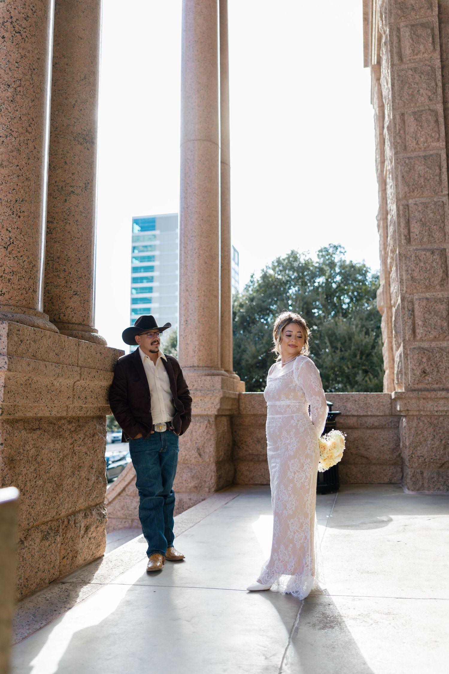 Bride in white lace gown holds bouquet beside groom in cowboy hat and jeans at a stone columned venue.