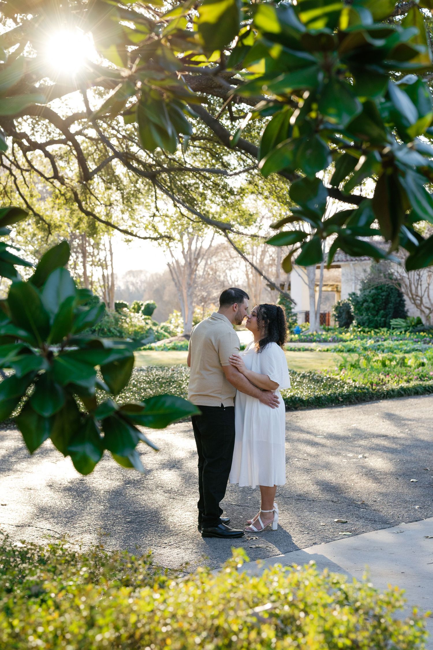 Couple sharing a romantic moment on a garden path, framed by lush magnolia leaves in golden sunlight.