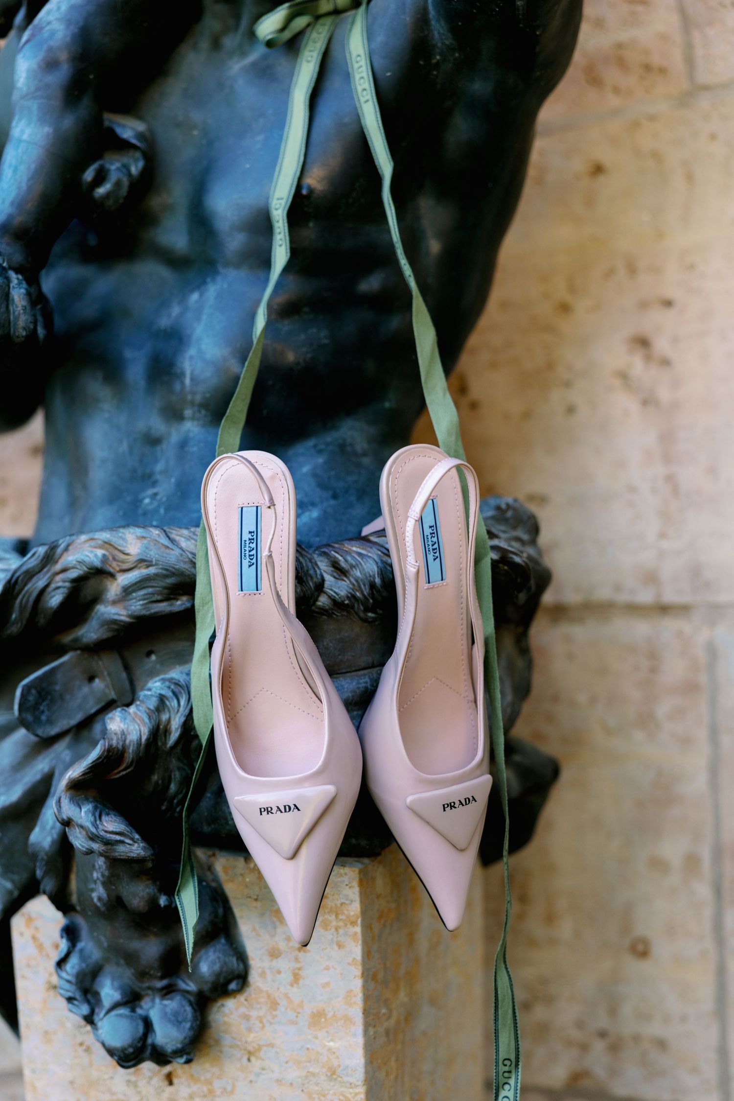 Blush pink Prada slingback heels resting on a bronze sculpture with green ribbon accents.