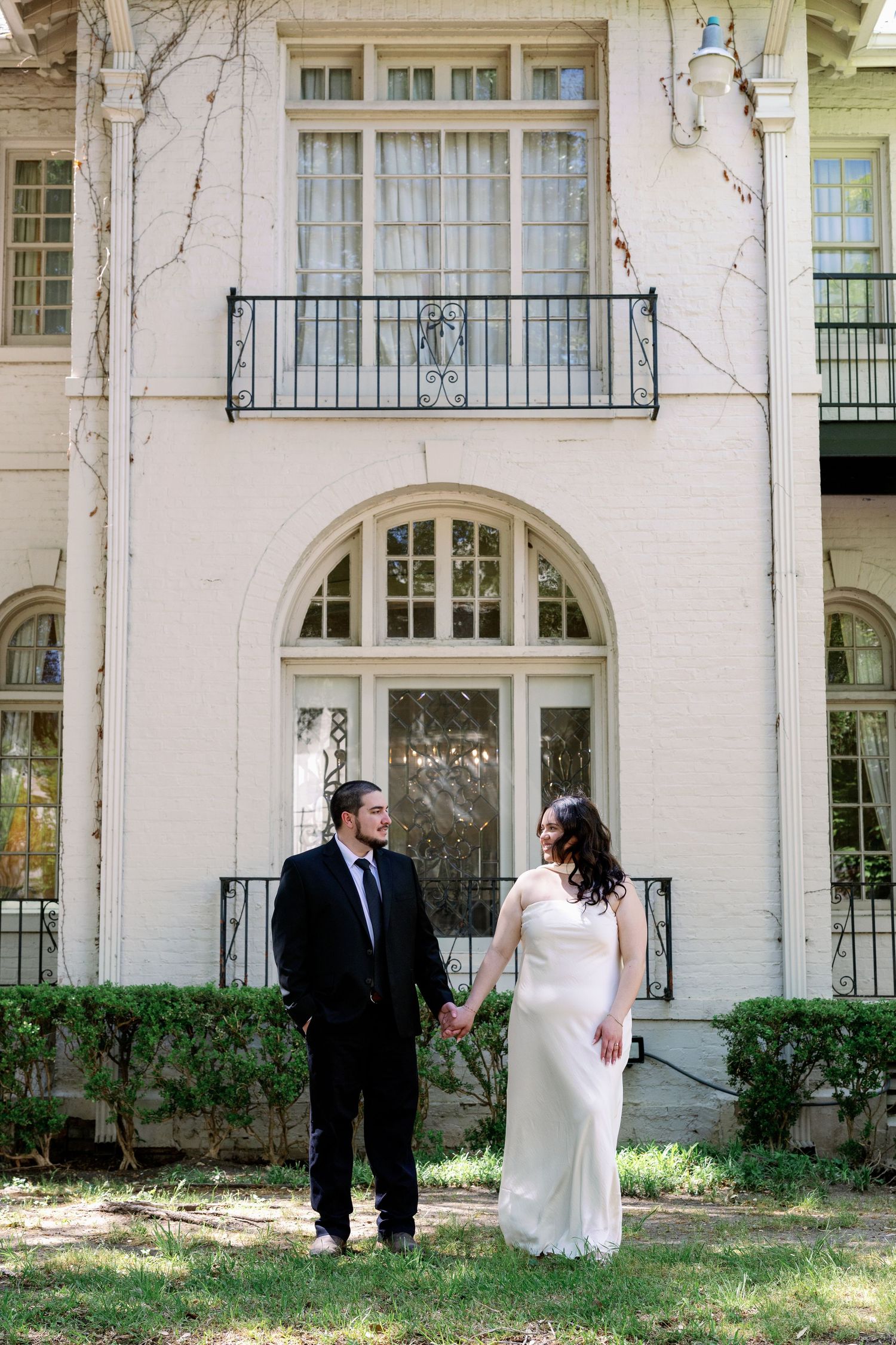 Bride and groom hold hands in front of elegant white brick mansion with arched windows and iron balcony.