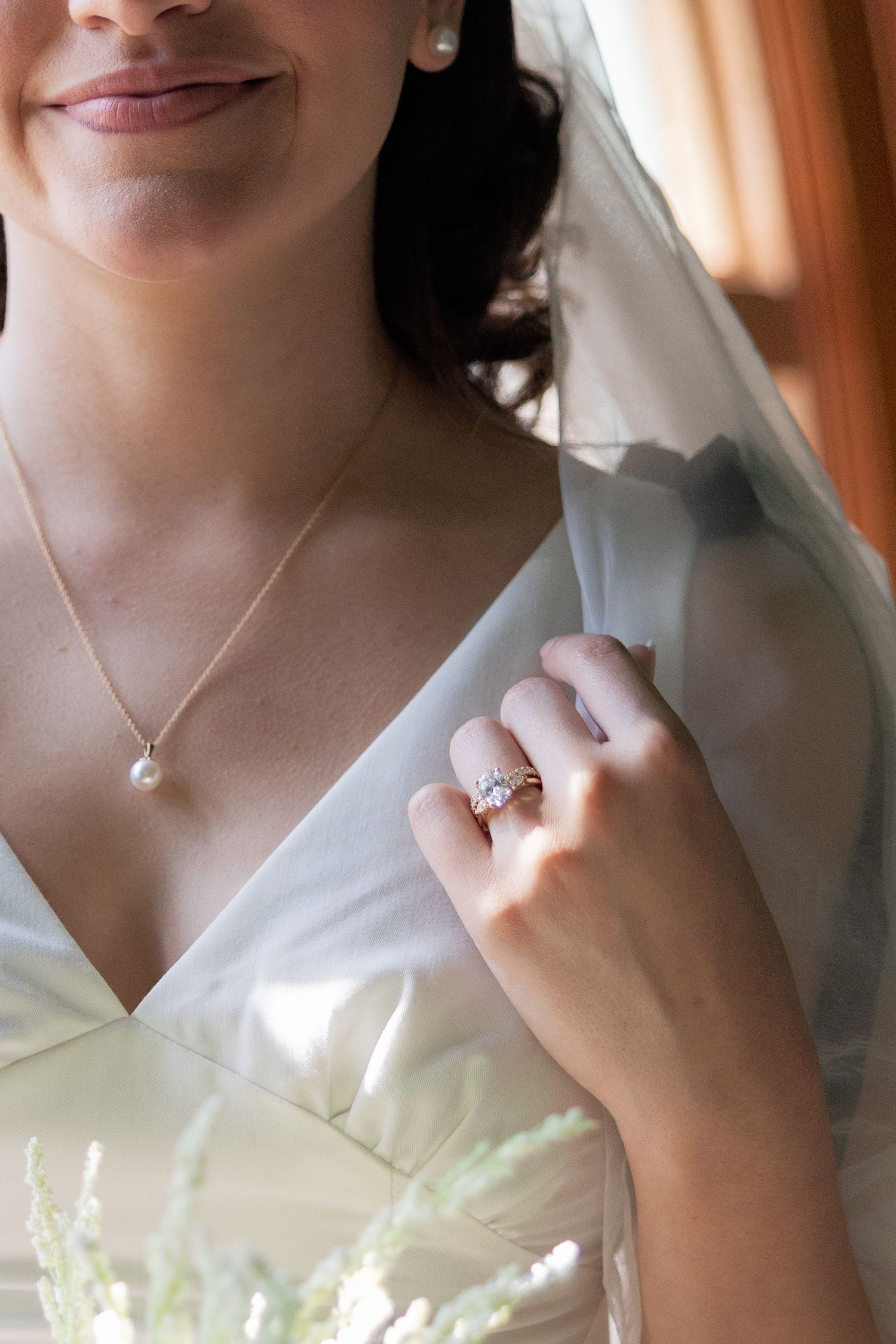 Bride in white gown holding veil, wearing diamond ring, pearl necklace, and stud earrings on wedding day.