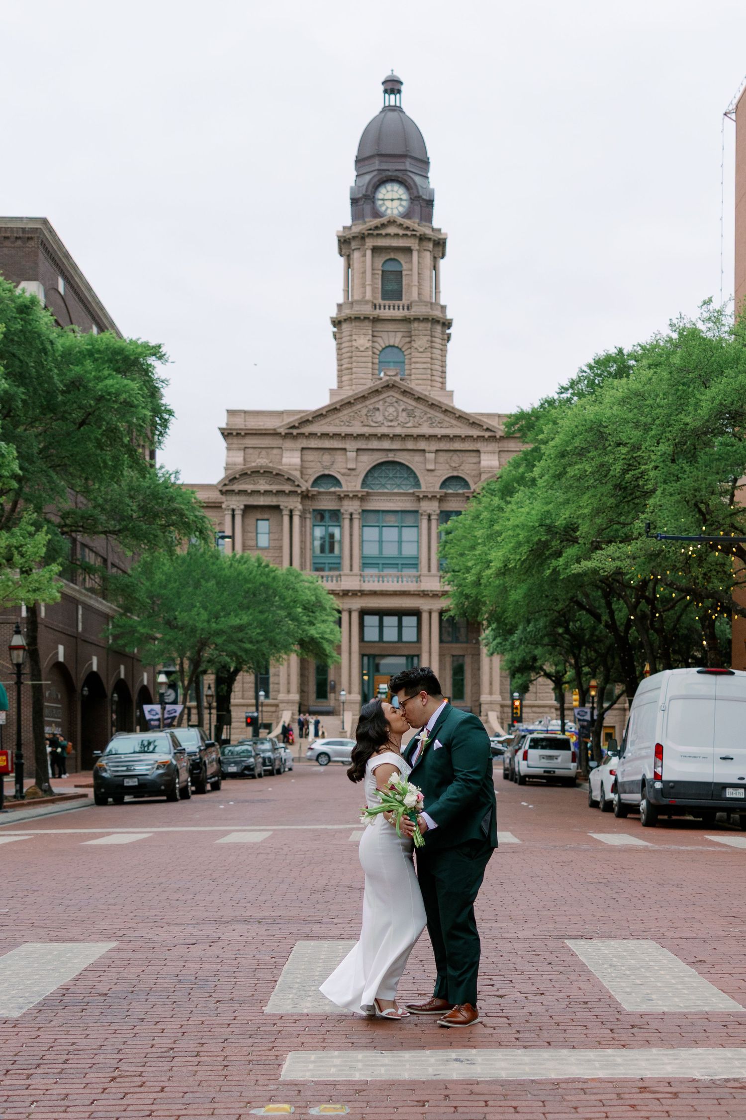 Bride and groom share a romantic kiss on a brick street in front of a historic courthouse with a clock tower.