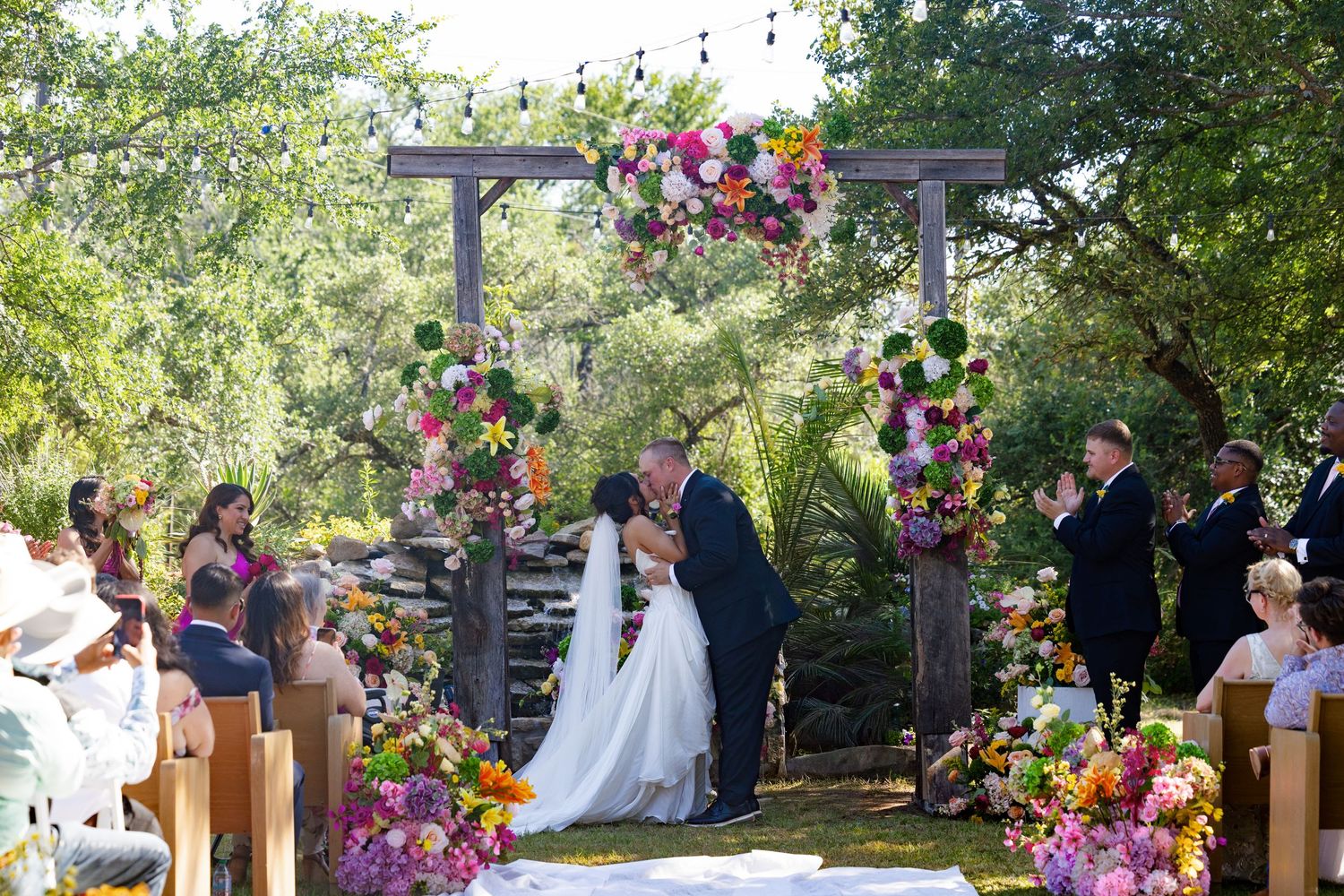 Couple shares first kiss under colorful floral arch at outdoor wedding ceremony with guests applauding.