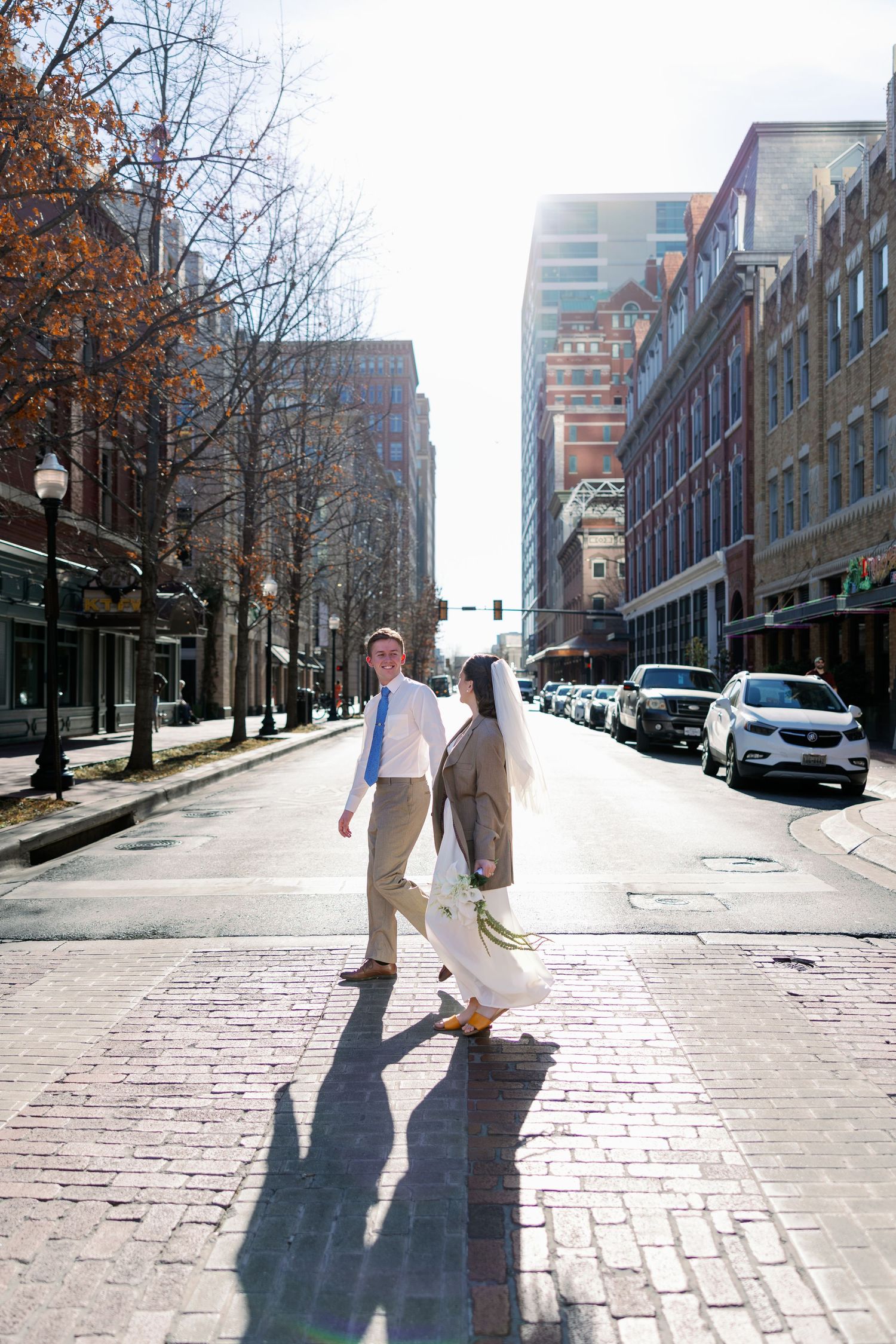 Bride and groom crossing a sunny brick street in an urban setting, surrounded by historic buildings and autumn trees.