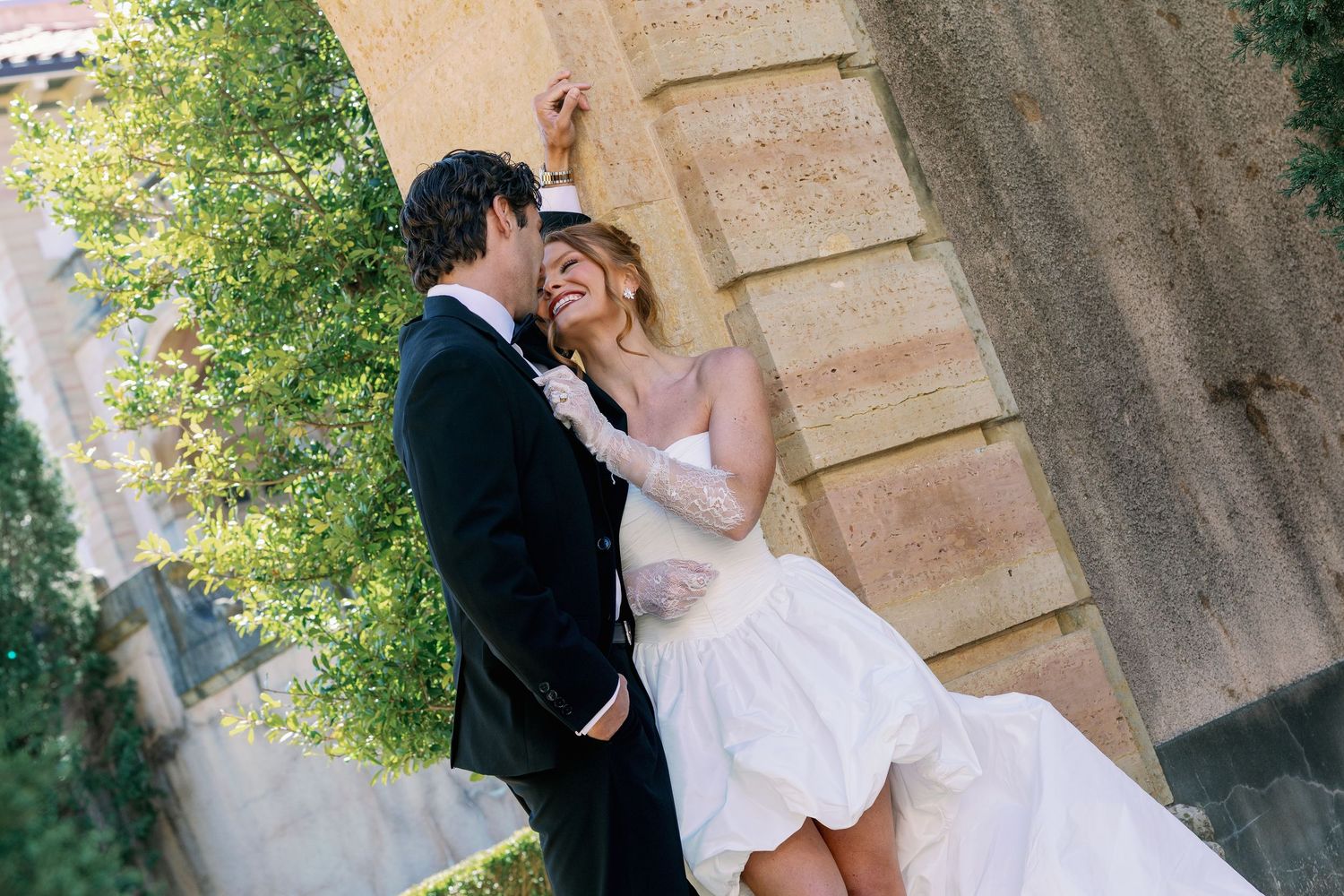 A joyful bride and groom share a romantic moment outdoors near a stone building on their wedding day.