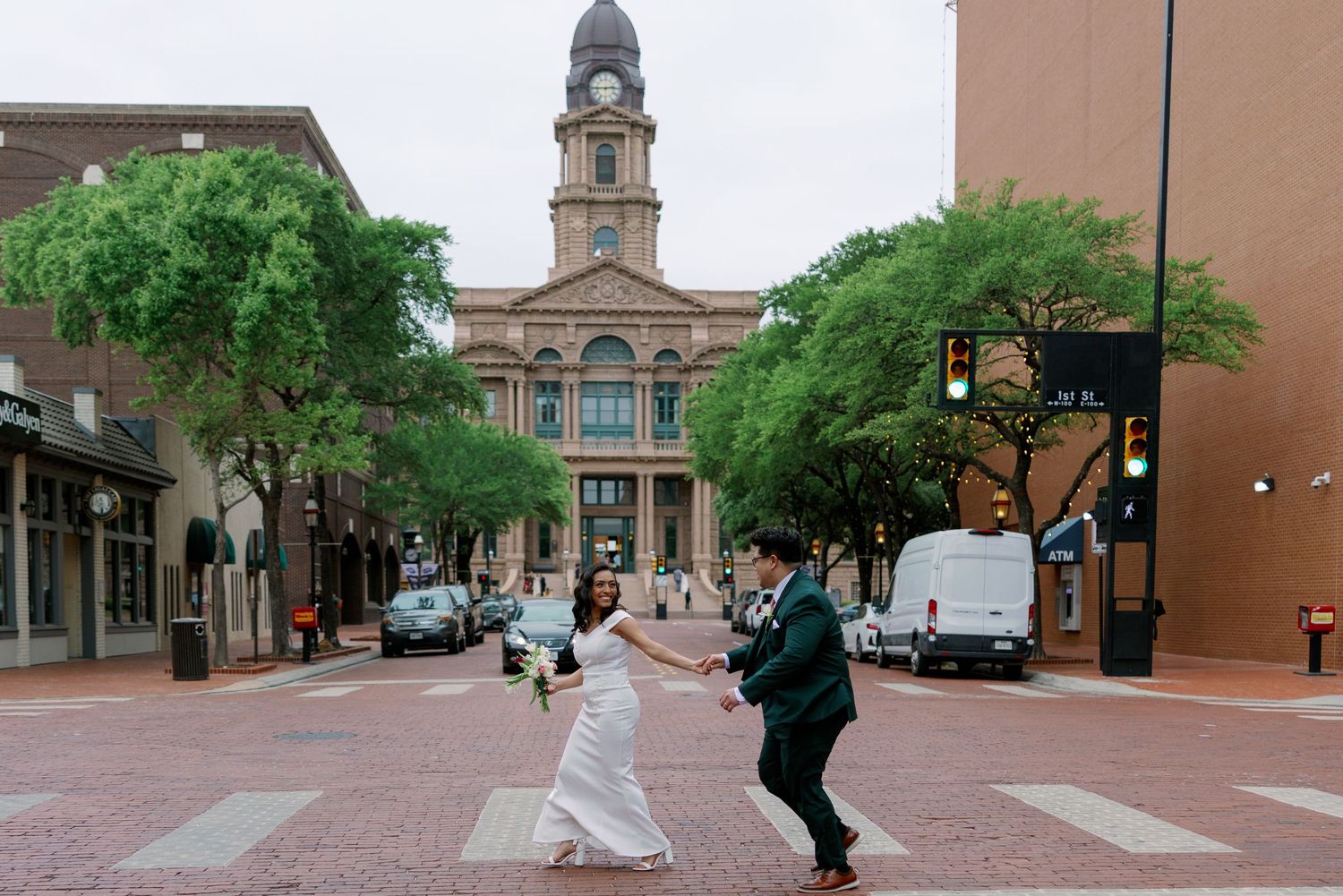 Bride and groom crossing a brick street holding hands in front of a historic courthouse in downtown Fort Worth, Texas.