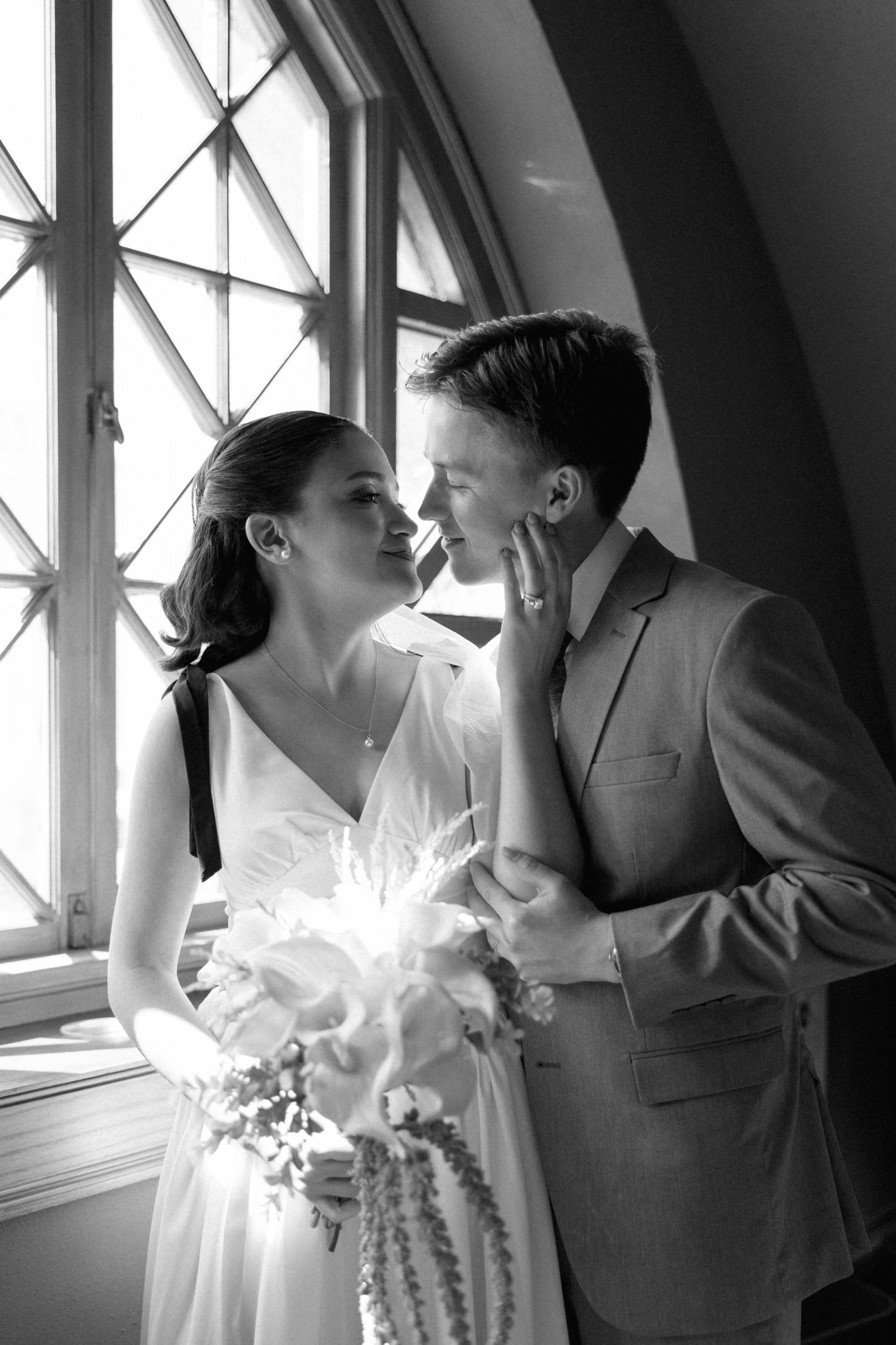 Black and white photo of a bride and groom sharing a romantic moment by a window, bride holding a bouquet.