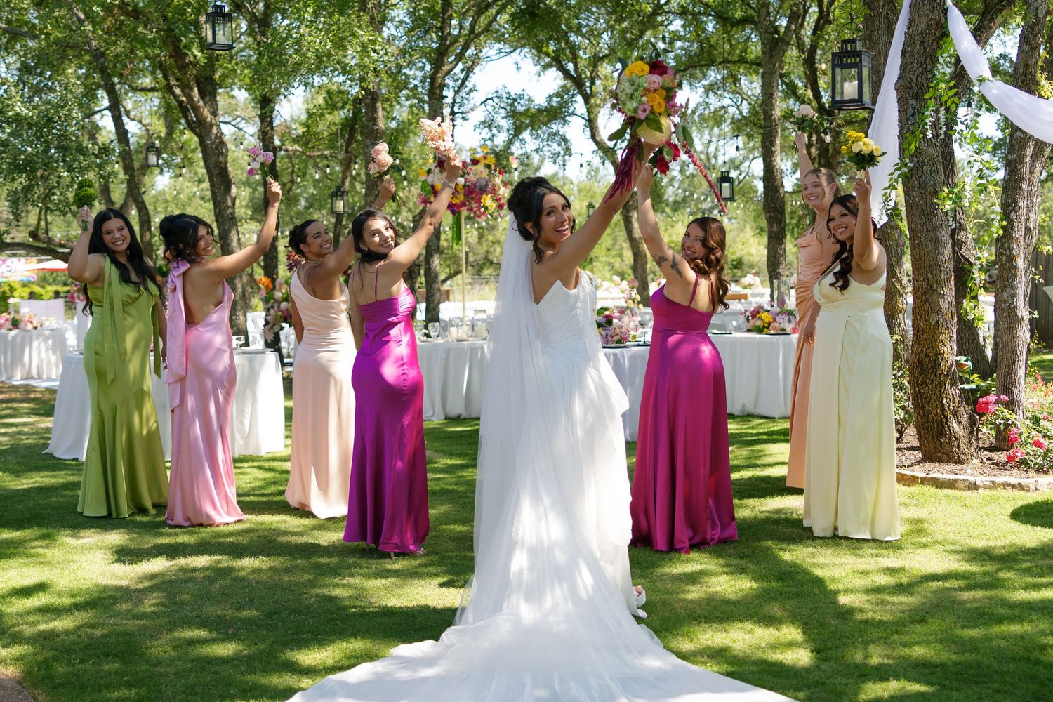 Bride in white gown celebrates with bridesmaids in colorful dresses raising bouquets outdoors at a garden wedding.