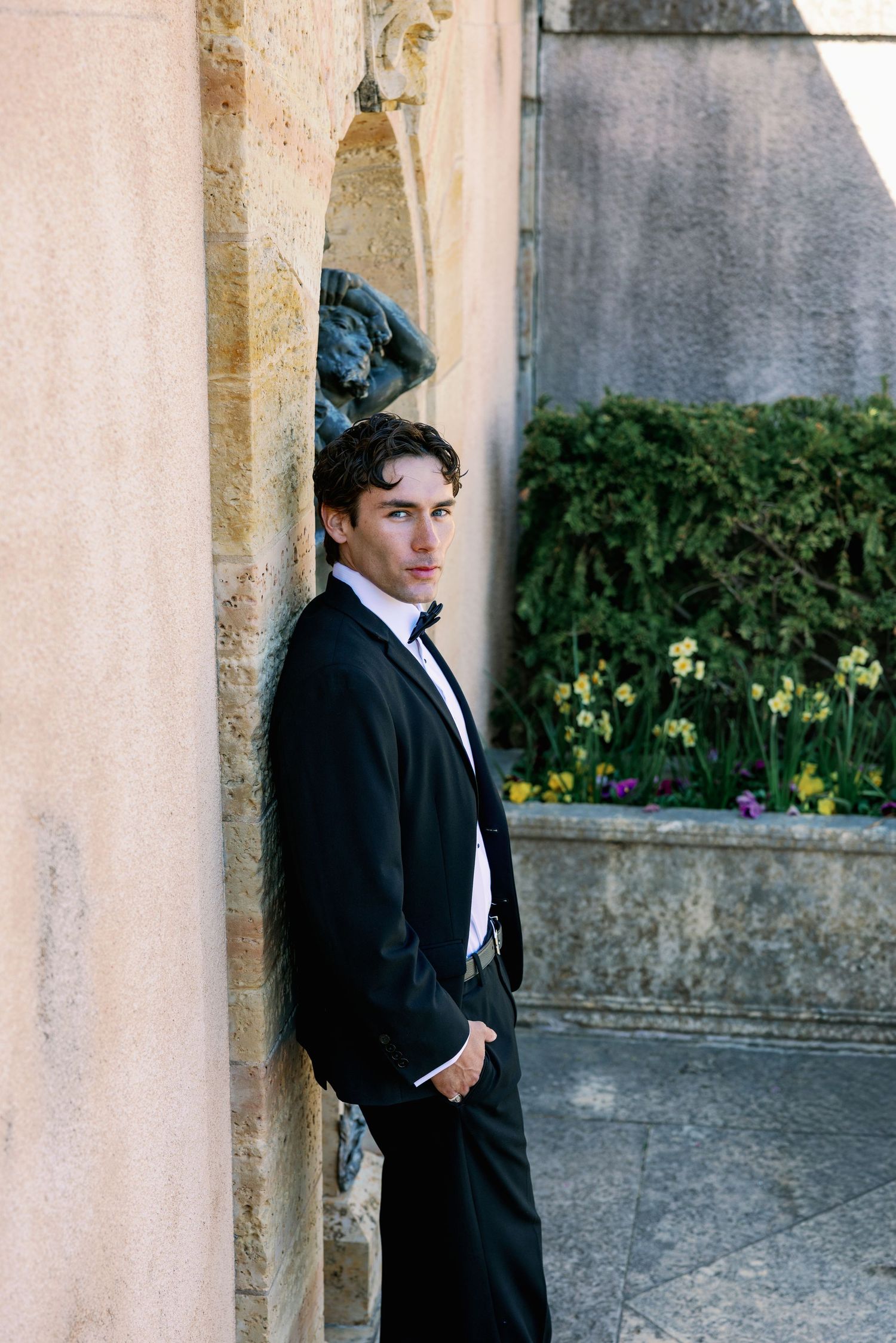 Young man in black tuxedo leaning against stone column outdoors with yellow flowers in background.