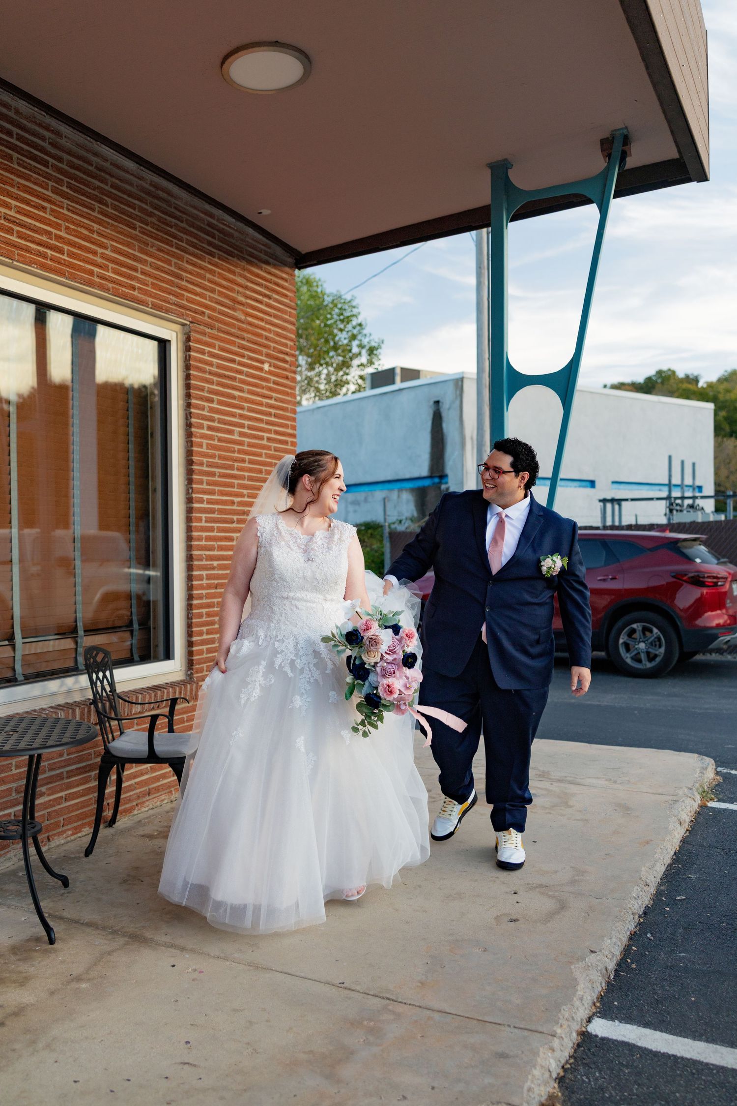 Bride and groom walking together outside a brick building, bride holding pink and blue bouquet in white gown.