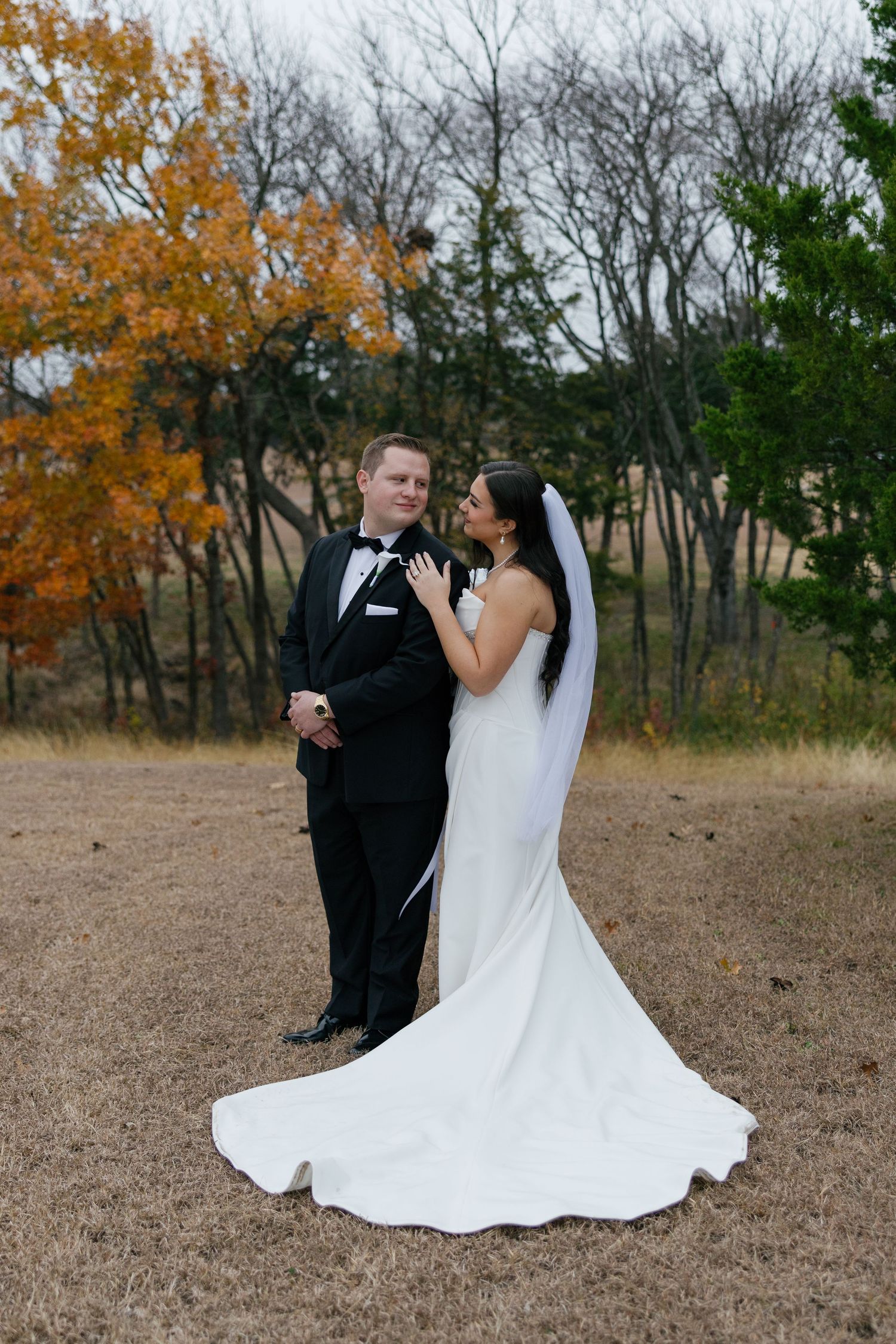 A couple in wedding attire sits together on dry grass with autumn trees creating a romantic fall backdrop.