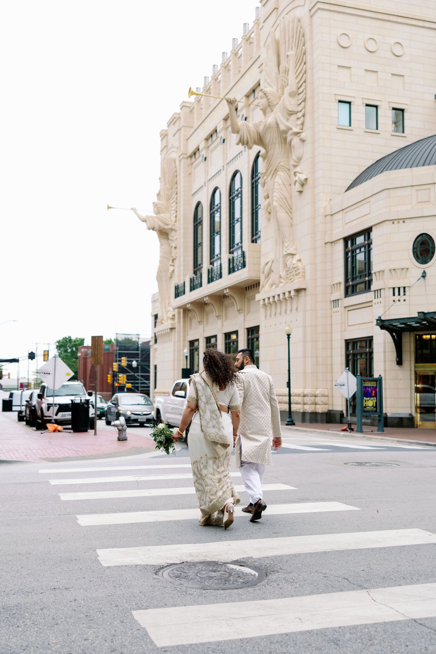 Couple in white wedding attire crossing a zebra crossing in front of an ornate beige building in the city.