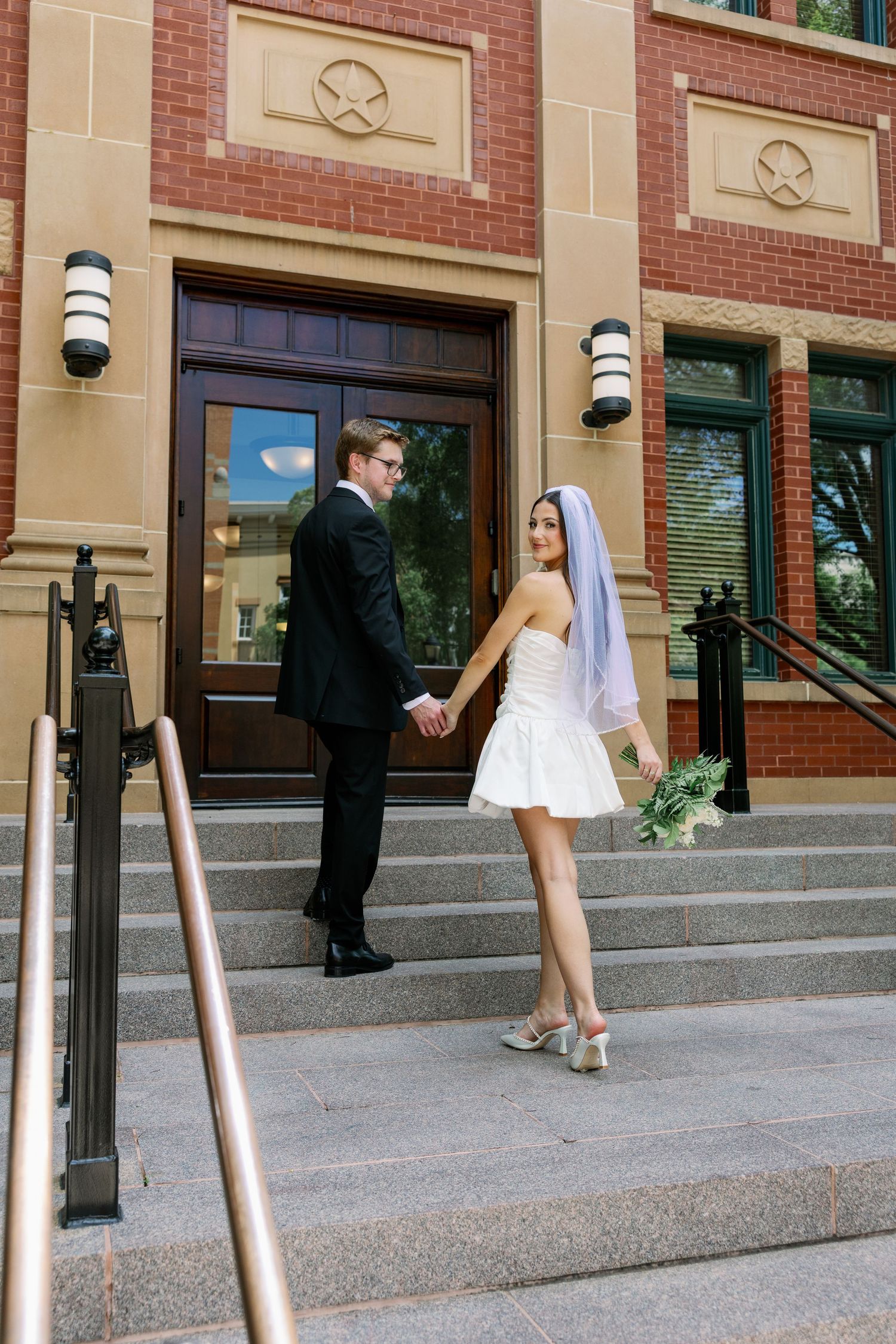 Bride in short white dress and veil holds groom's hand on steps of brick building during outdoor wedding photo.
