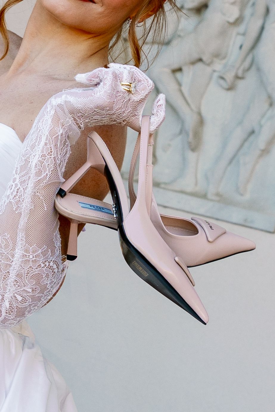 Bride in lace wedding dress holding blush pink Prada slingback heels outdoors.