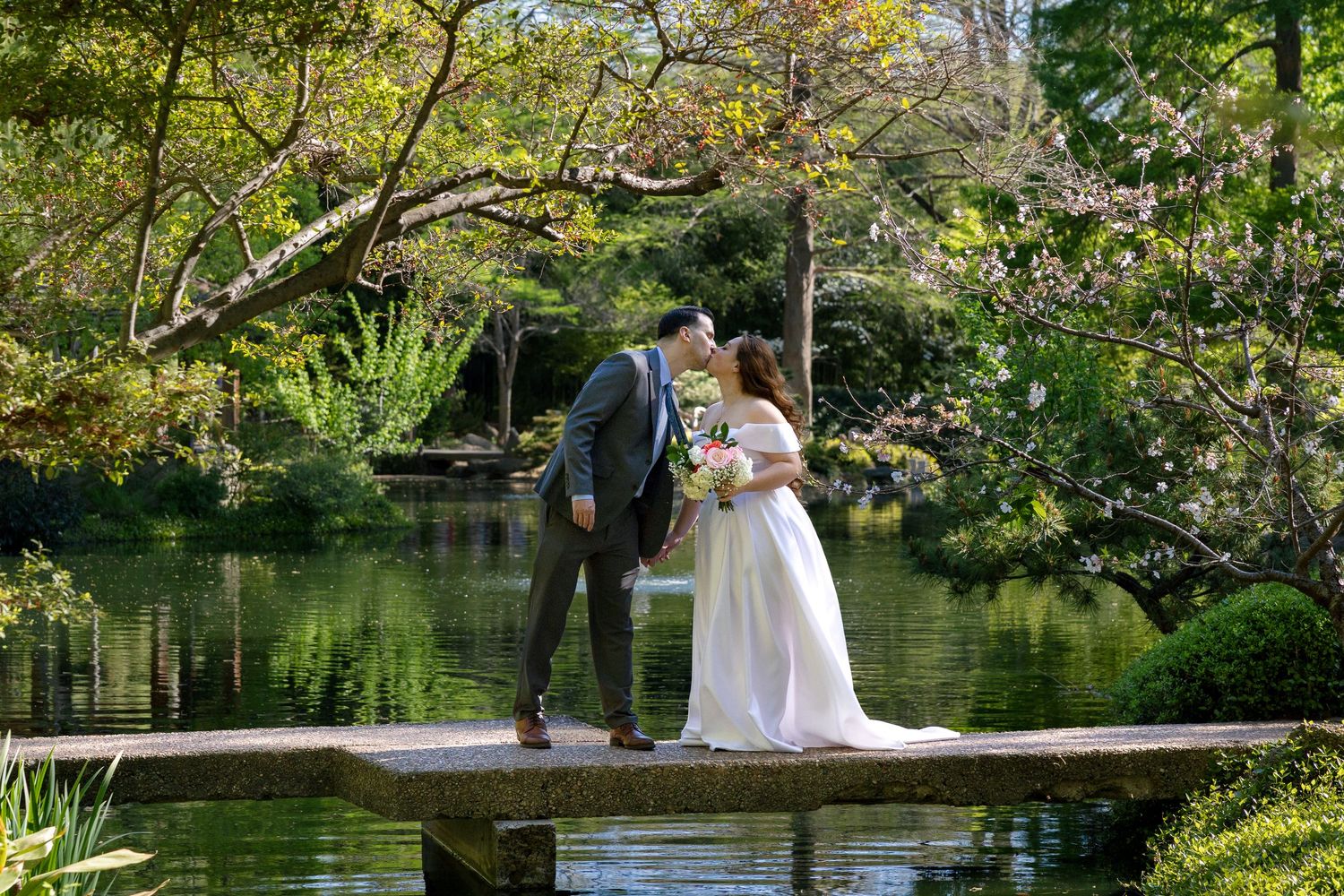 Bride and groom share a kiss on a stone pier overlooking a serene garden pond surrounded by lush green trees.