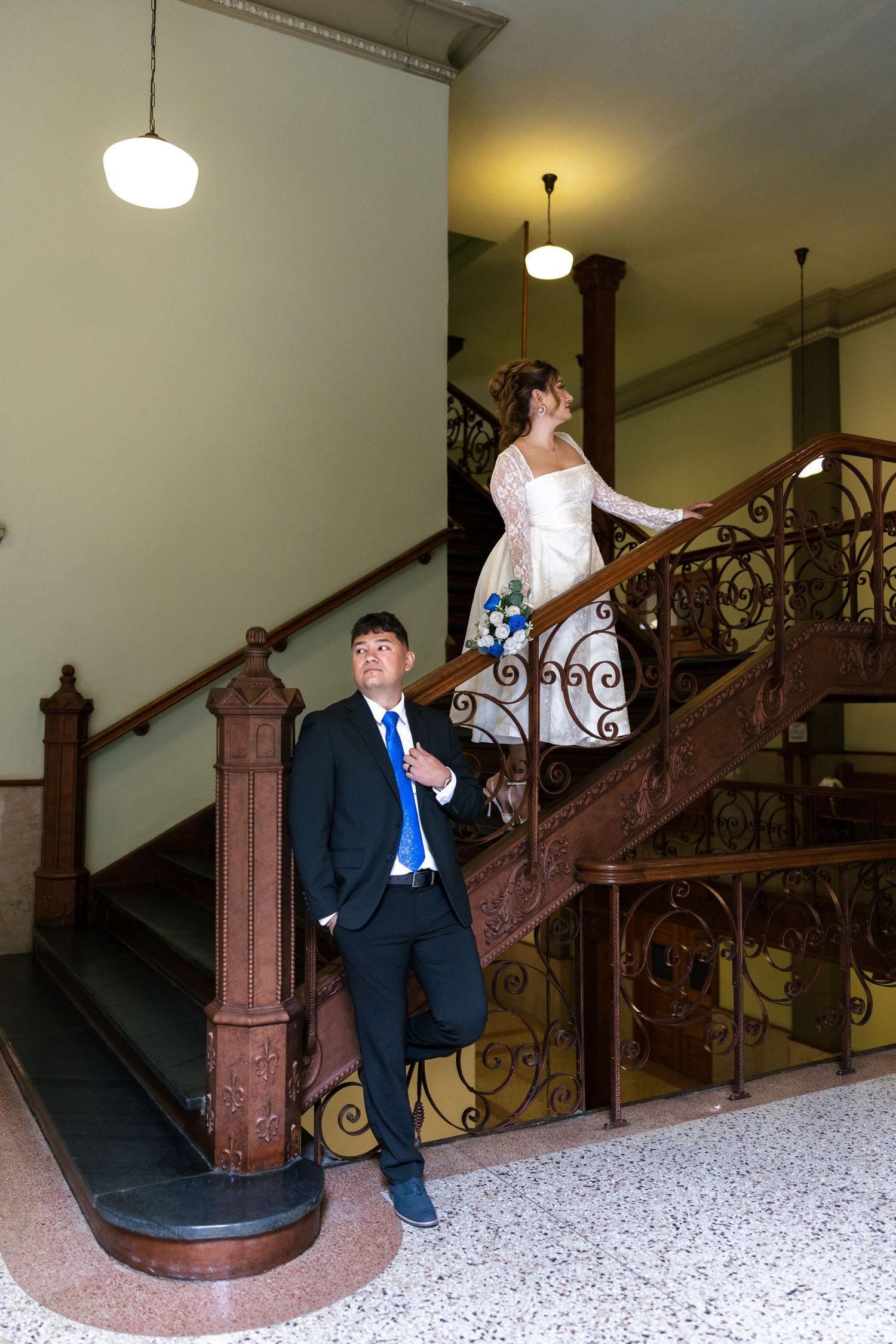 Bride in white gown descends ornate wooden staircase while groom in black suit with blue tie waits below in elegant venue.
