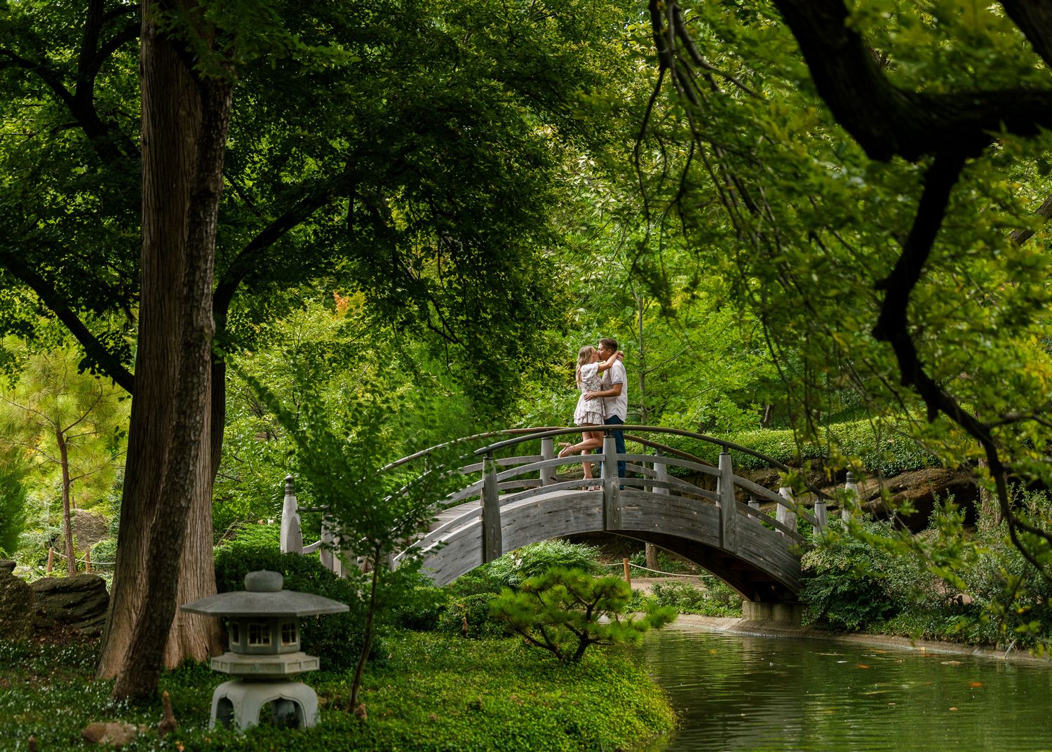 Romantic couple walking along scenic garden paths with lush green trees and stone bridges.